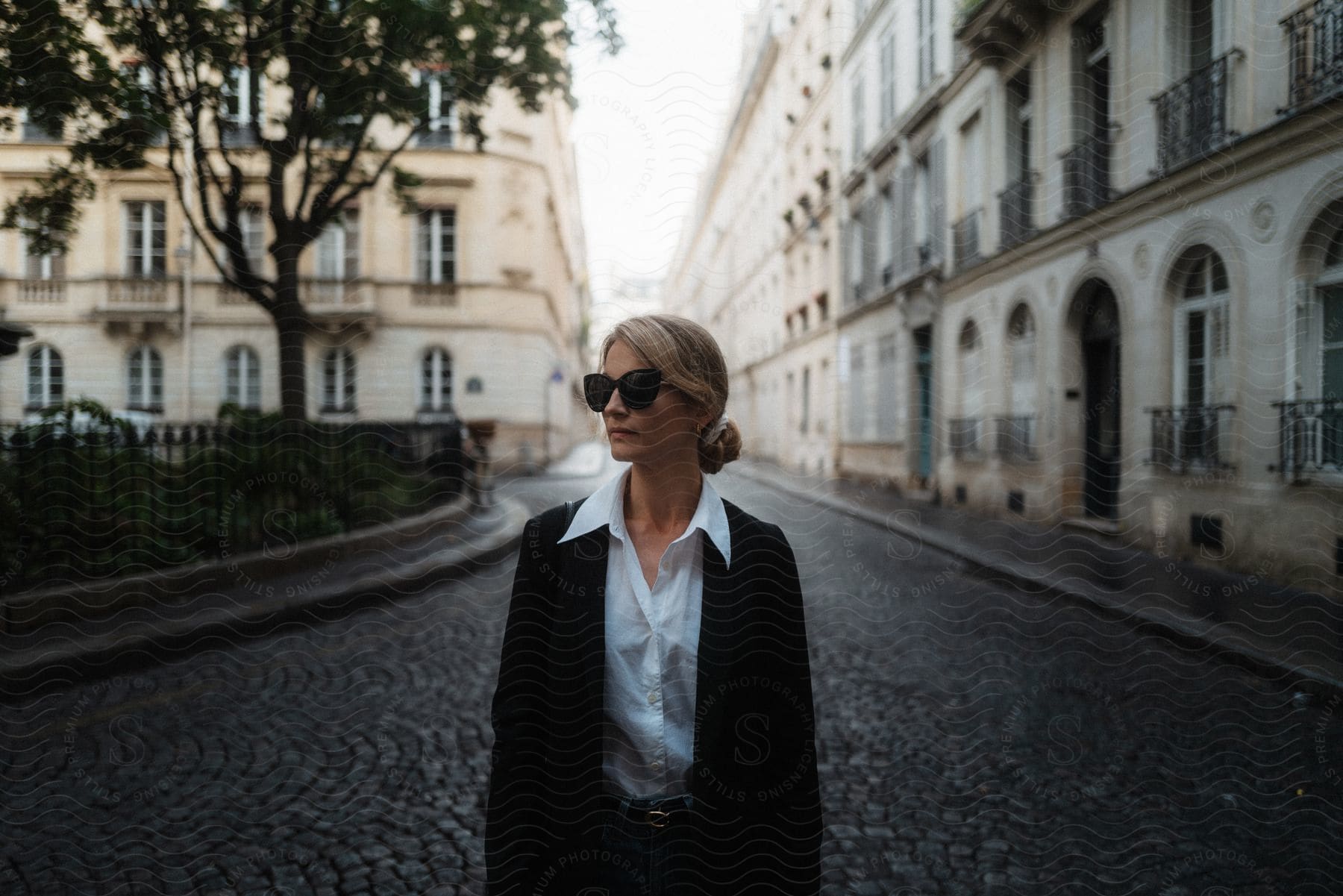 Blonde woman wearing sunglasses and black jacket stands in cobblestone roundabout with buildings trees and bushes