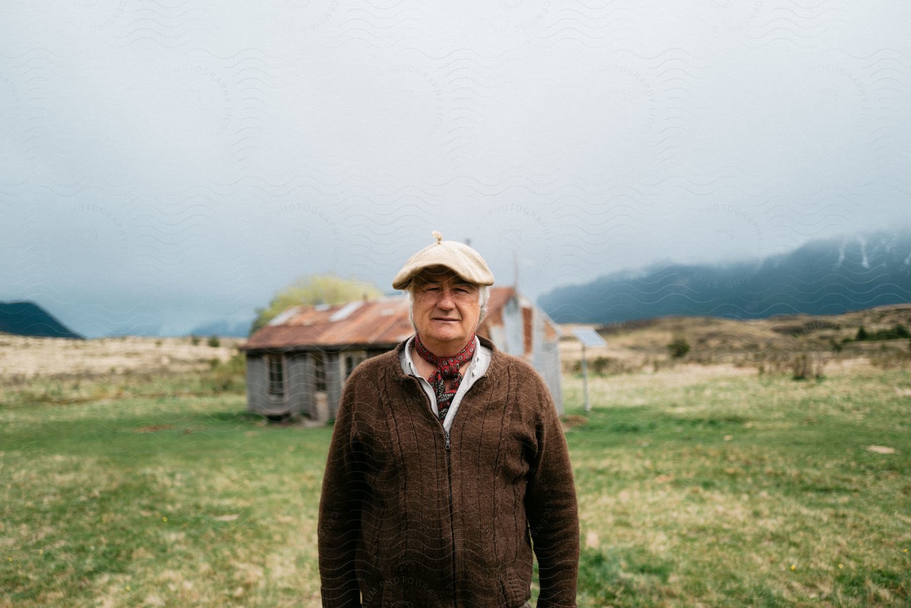 Elderly man in beret stands in front of farmhouse with snowy mountains in the background