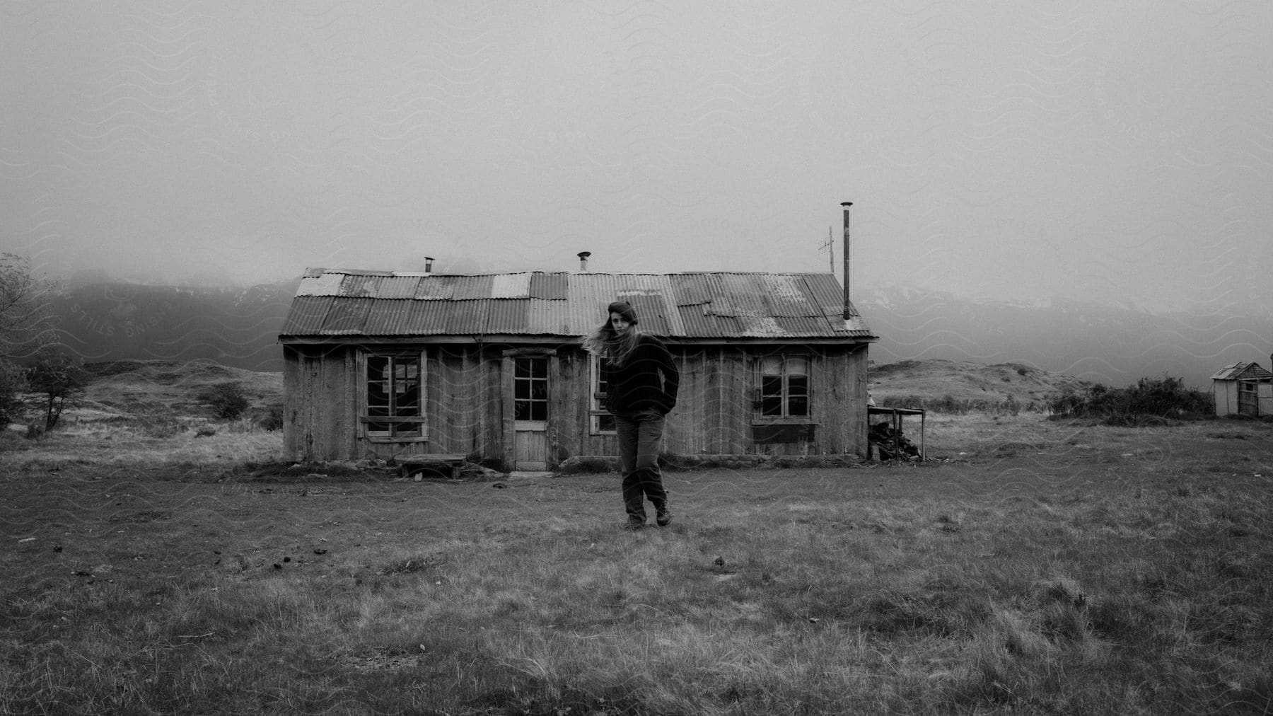 A woman with windblown hair stands in front of a cabin in a field on a foggy day