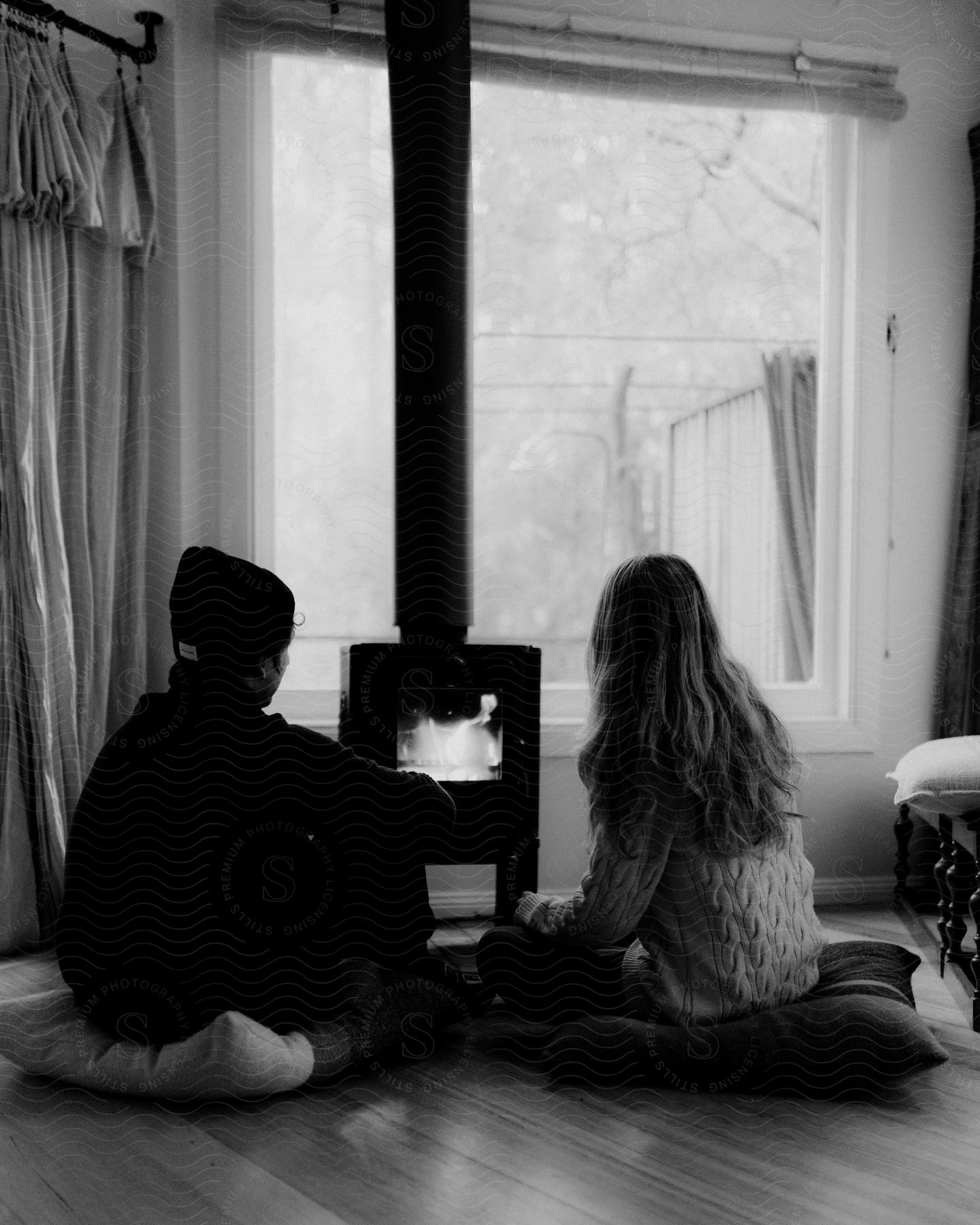 A man and woman sit on the floor near a wood burning stove in an interior setting