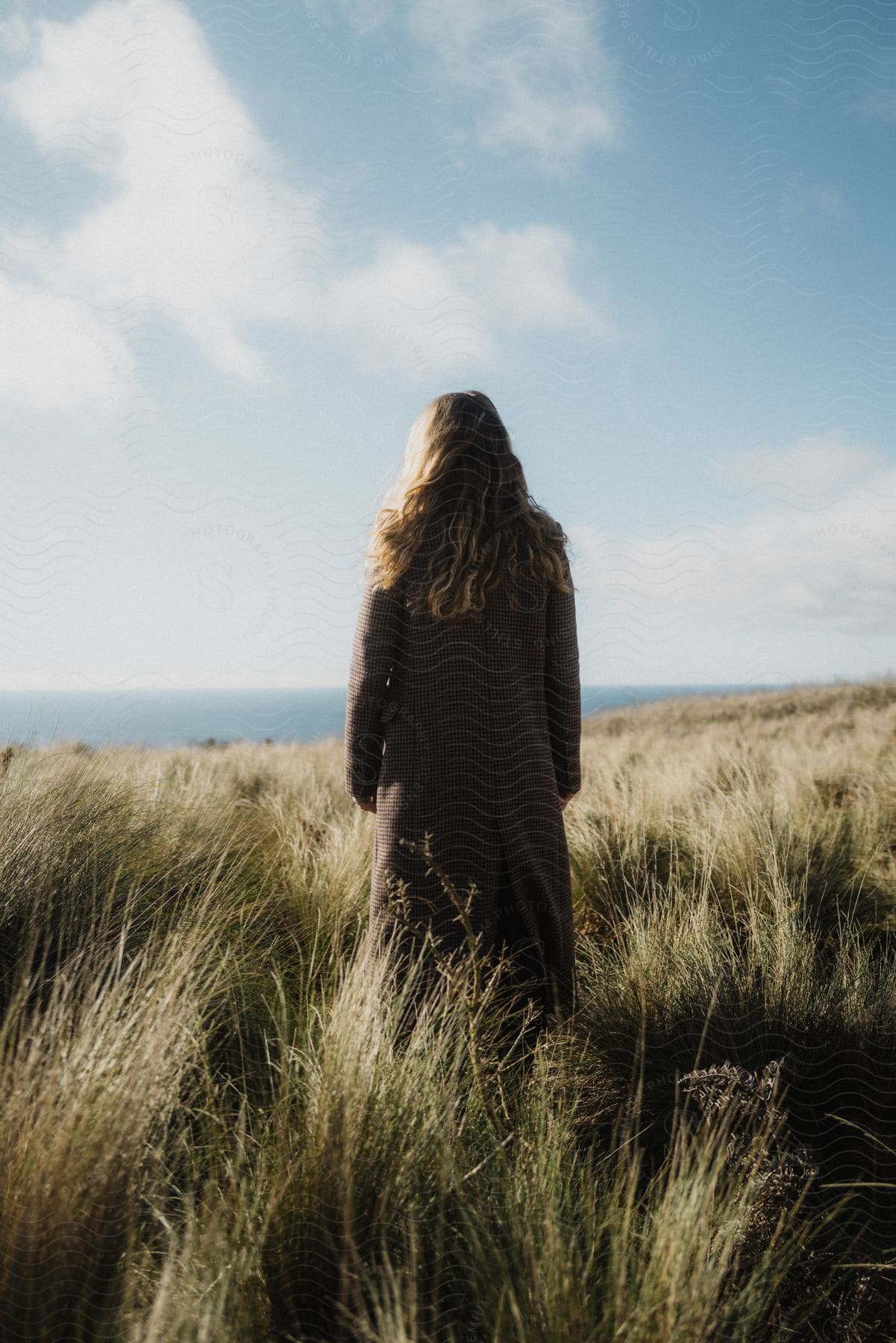 Woman posing in tall grass with long coat with coastal landscape in background