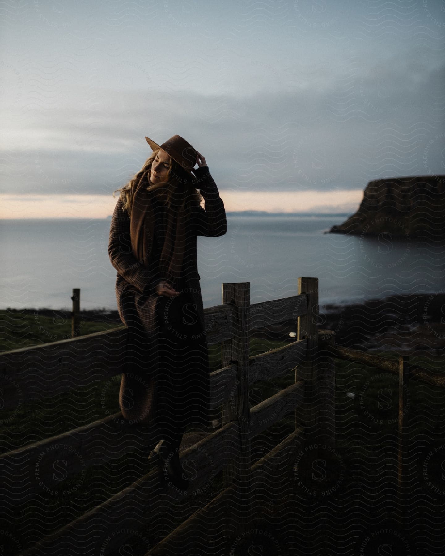 A woman sitting on a wooden fence near the beach wearing a hat