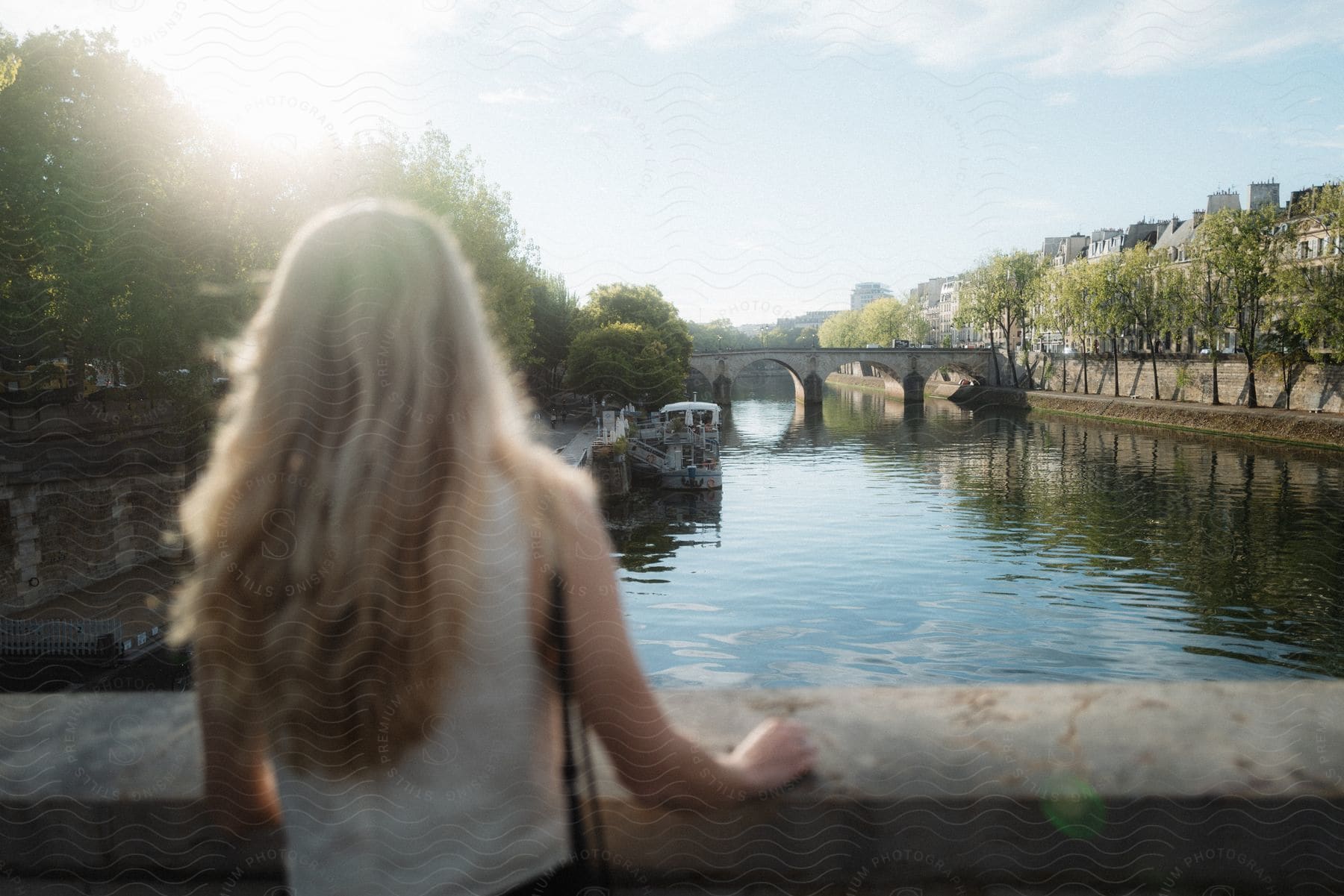 A woman on a bridge gazes at a boat on a river in a european city