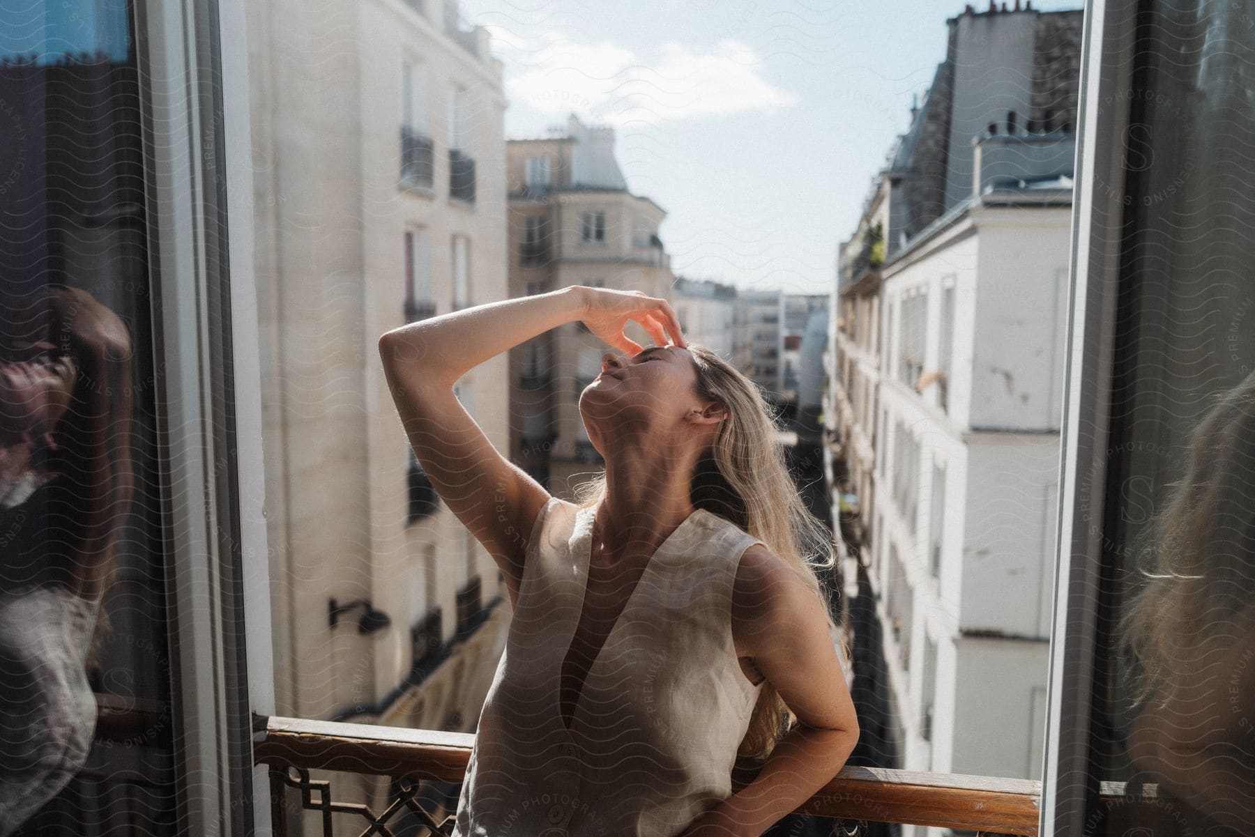 A woman standing near a window with her head raised and her hand on her forehead