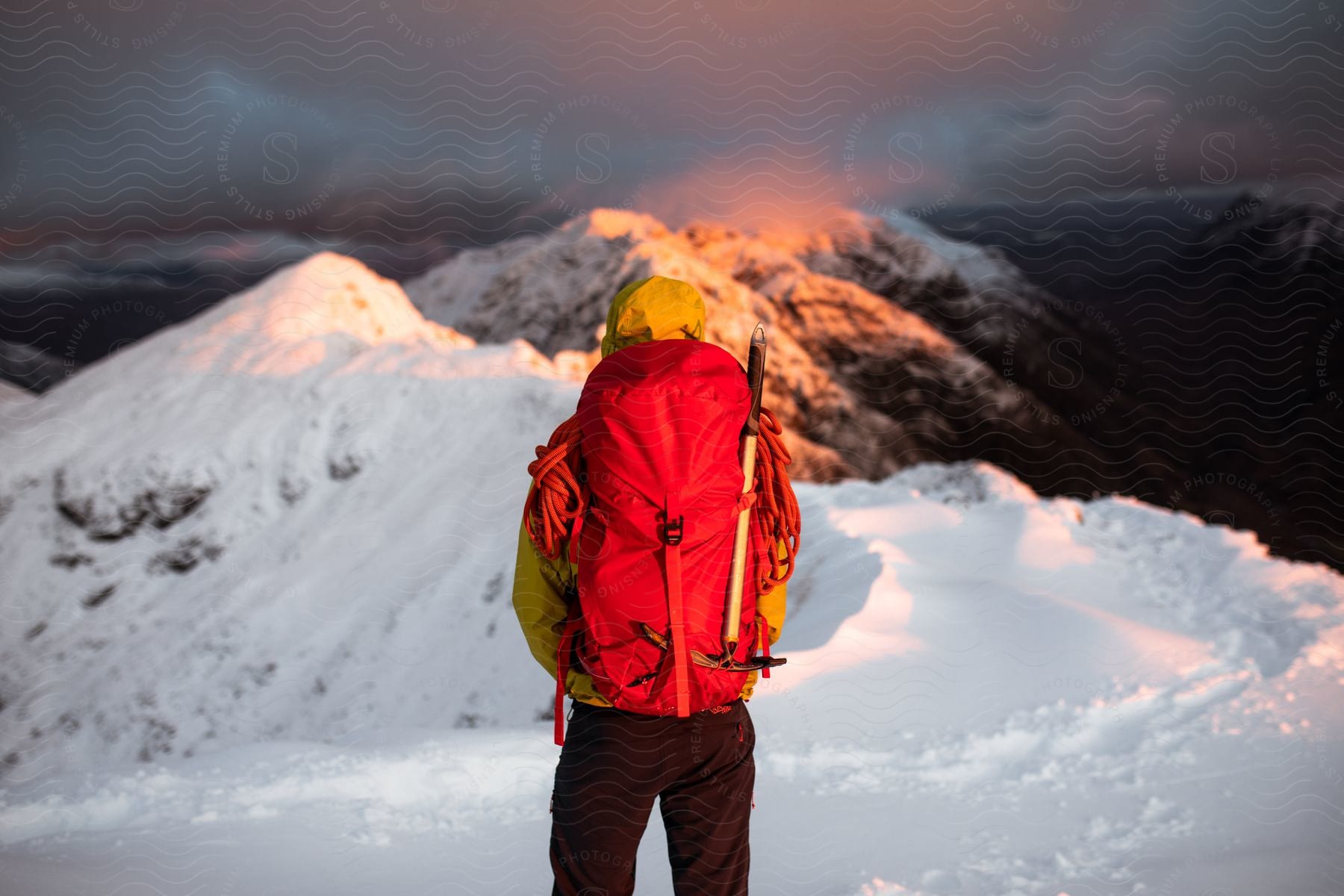 Man in yellow coat and red backpack observes snowy mountains