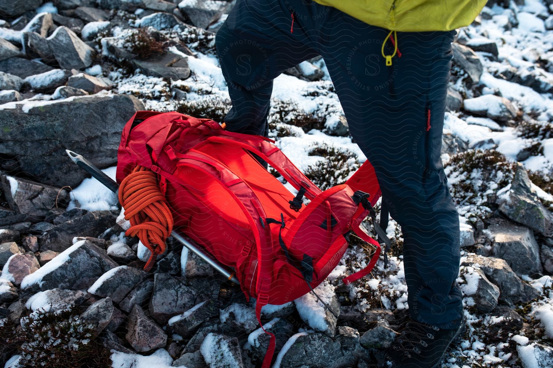 Man in ski pants and yellow coat stands by red backpack lying on rocky and snowy mountain slope