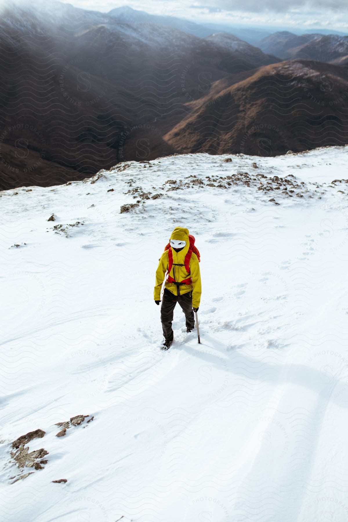 A hiker walks up a snowy path surrounded by mountains wearing a yellow coat and red backpack