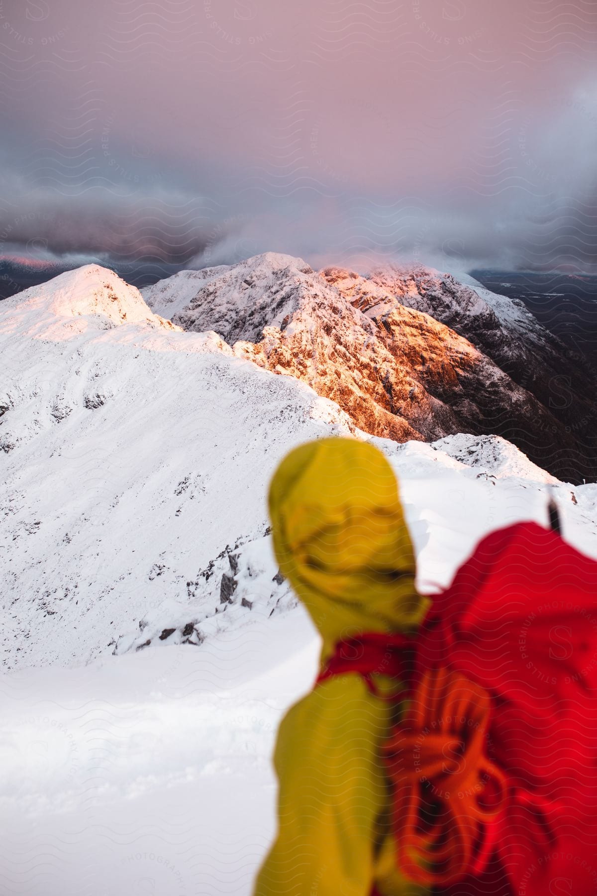Hiker with red backpack and yellow jacket looks out over snowy mountains
