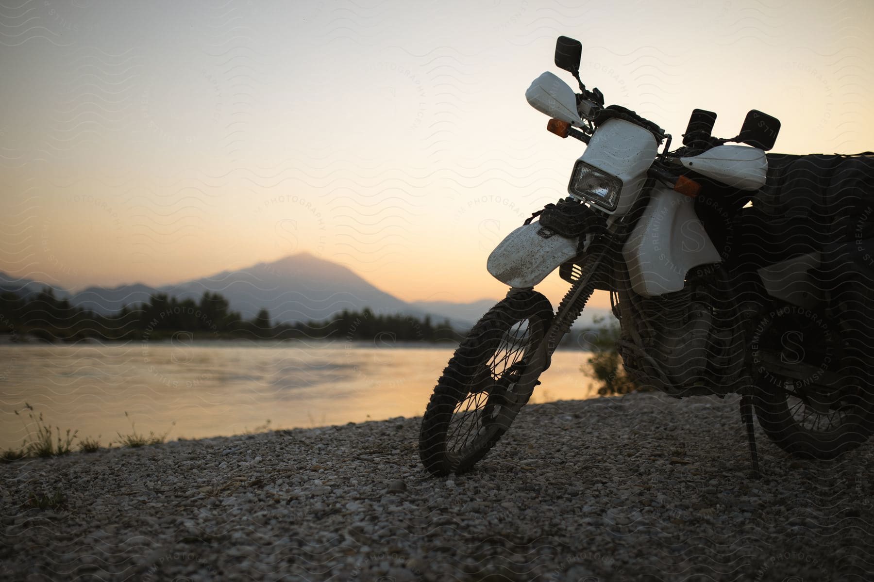 A white motorcycle stands along the shore with trees and a mountain across the river under a hazy sky