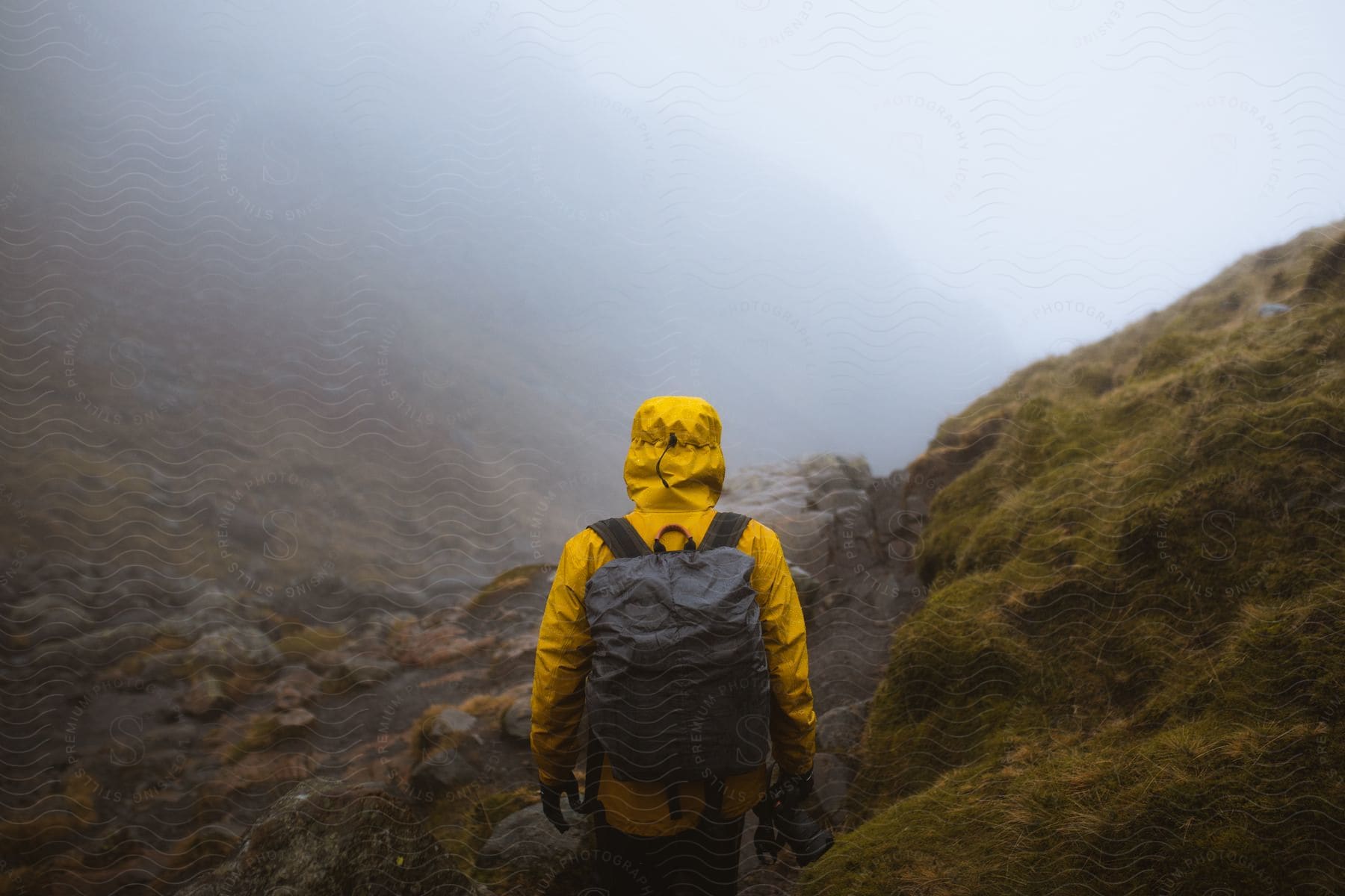 A person walking alone on rocky terrain with grasses carrying a bag and wearing a yellow jacket