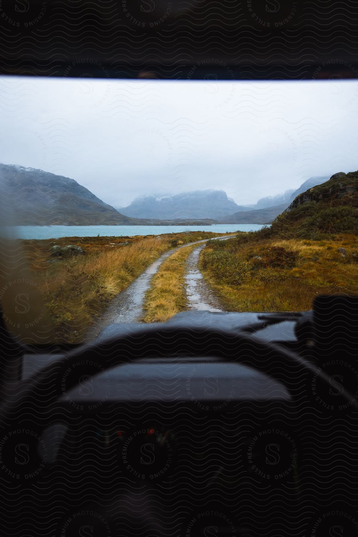 A car drives along a wet country road near a body of water and mountains in cloudy weather