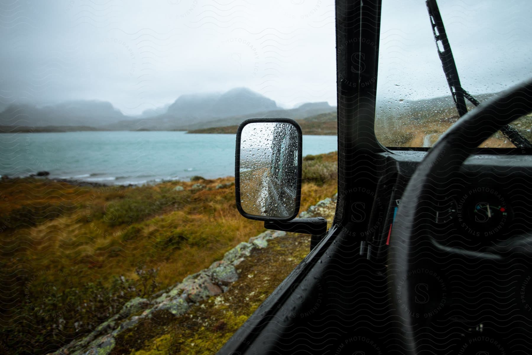 Raindrops cover the cars side view mirror and windshield as a serene lake is surrounded by rocks and grass