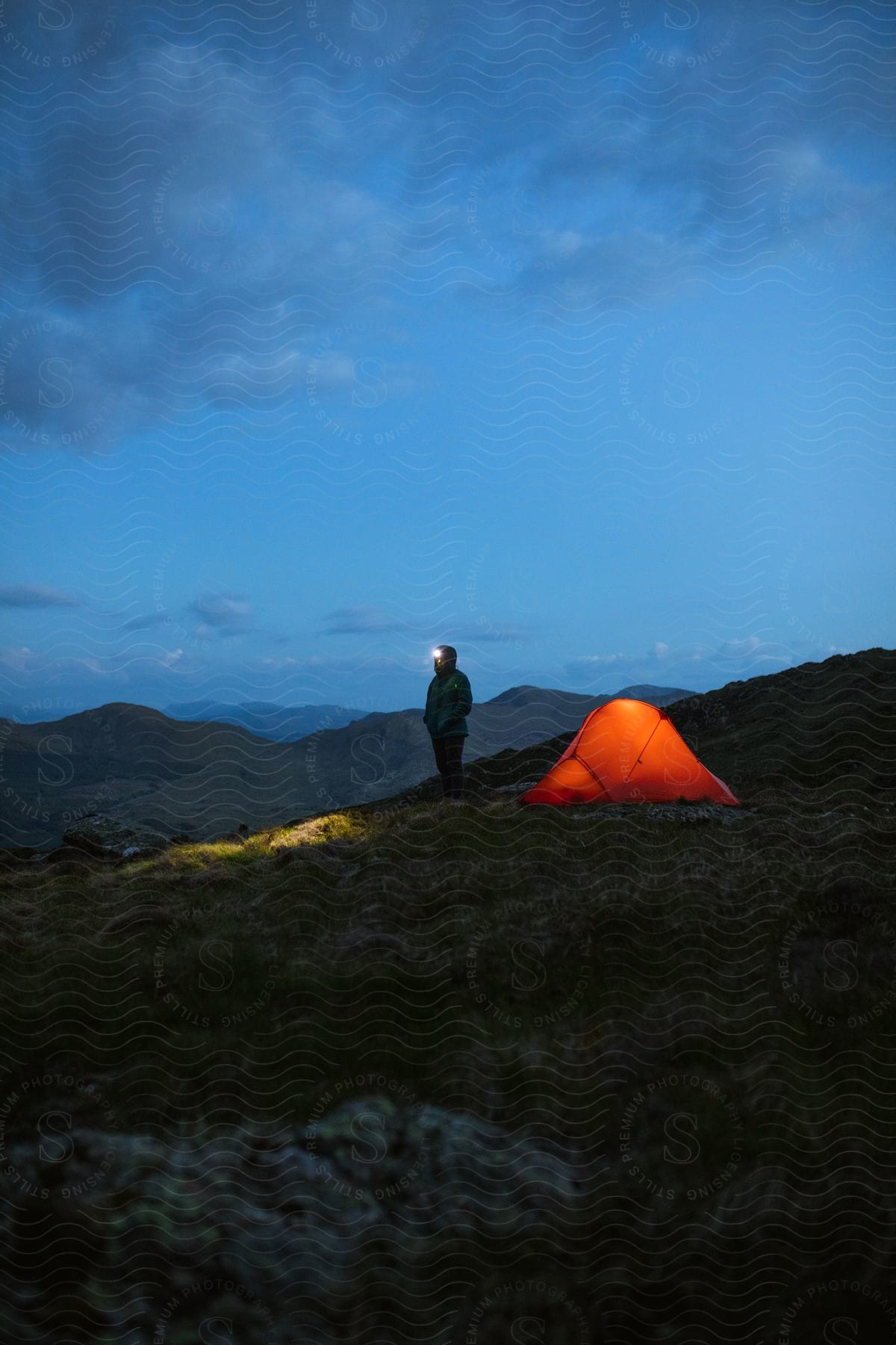 A camper stands by his red tent at night wearing a headlight in a campsite nestled between rocky grassy hills under a cloudy sky