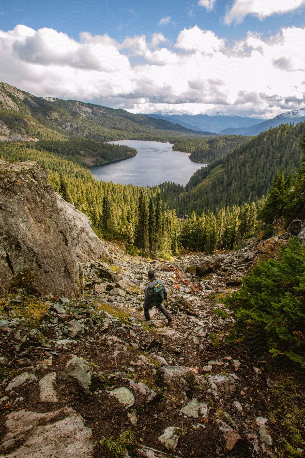 A man walks over a rock formation with a forest and lake in front of him in the distance