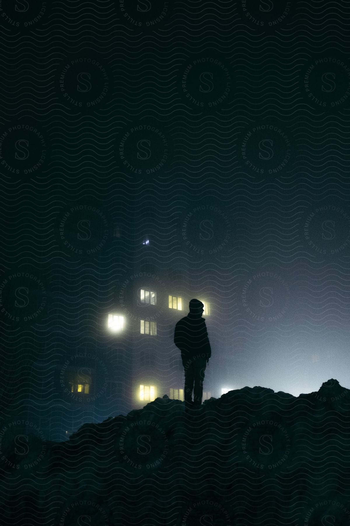 A man silhouetted on a rubble pile with a lit building in the background at night