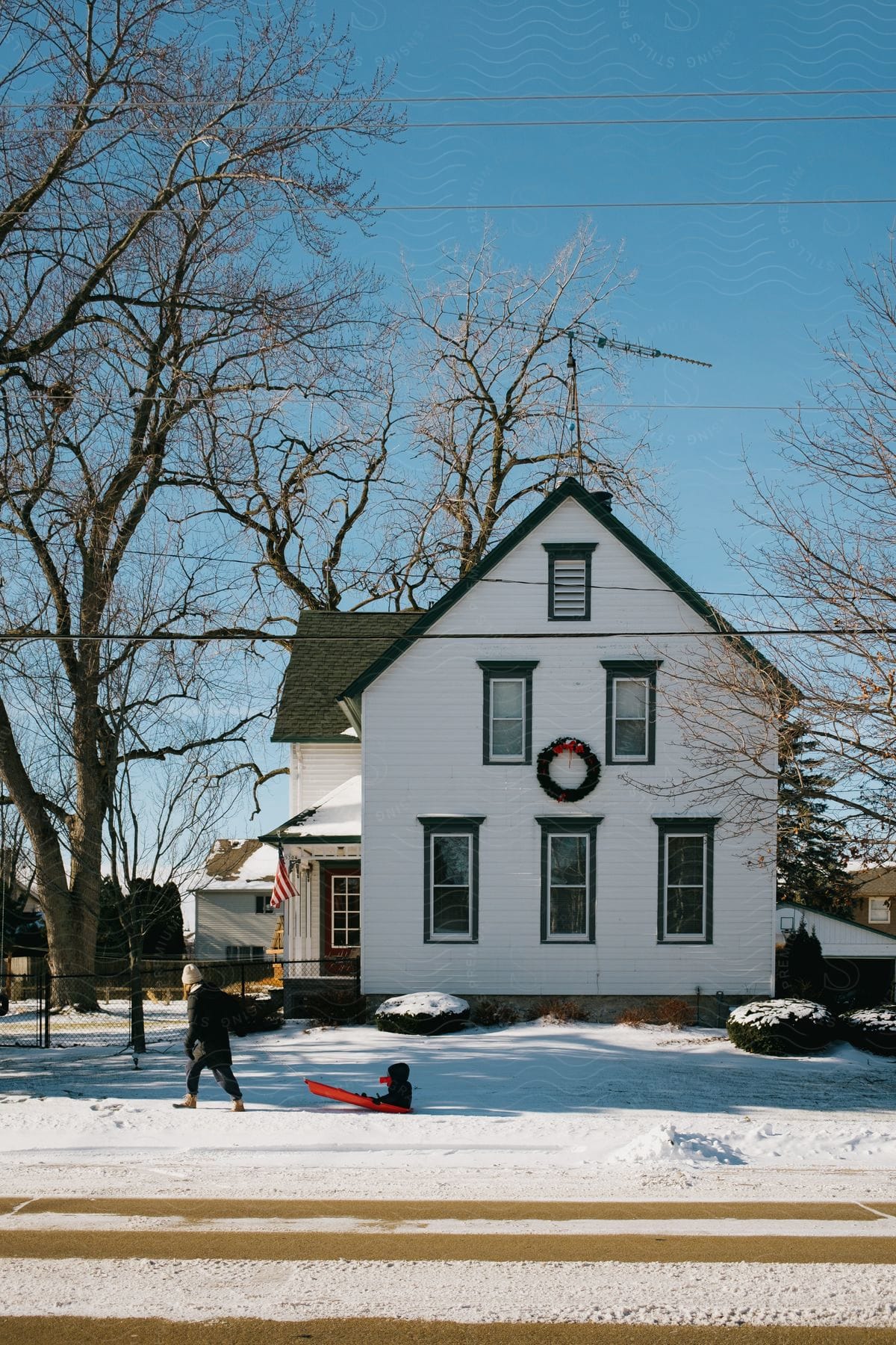 Adult walking down snowcovered sidewalk pulling child on sled in front of twostory white home on winter day