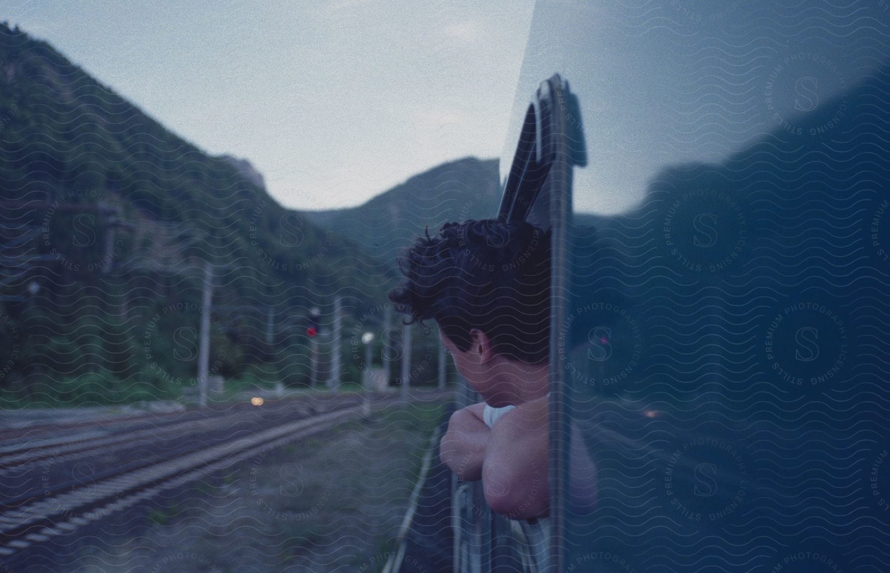 A man leans out of a train window as it travels through a mountainous region on a cloudy day