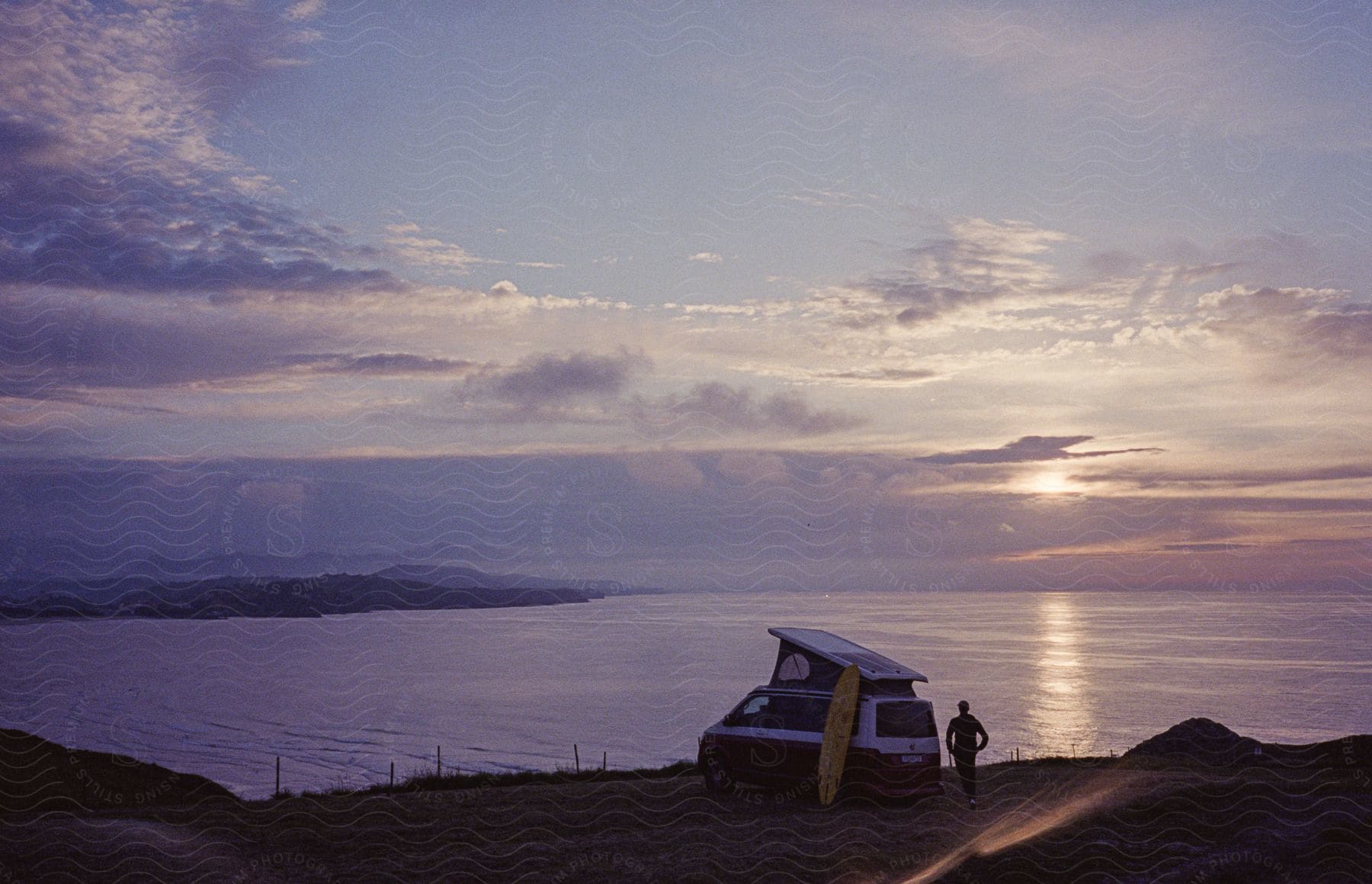A man stands next to his car near the shoreline at dusk or dawn