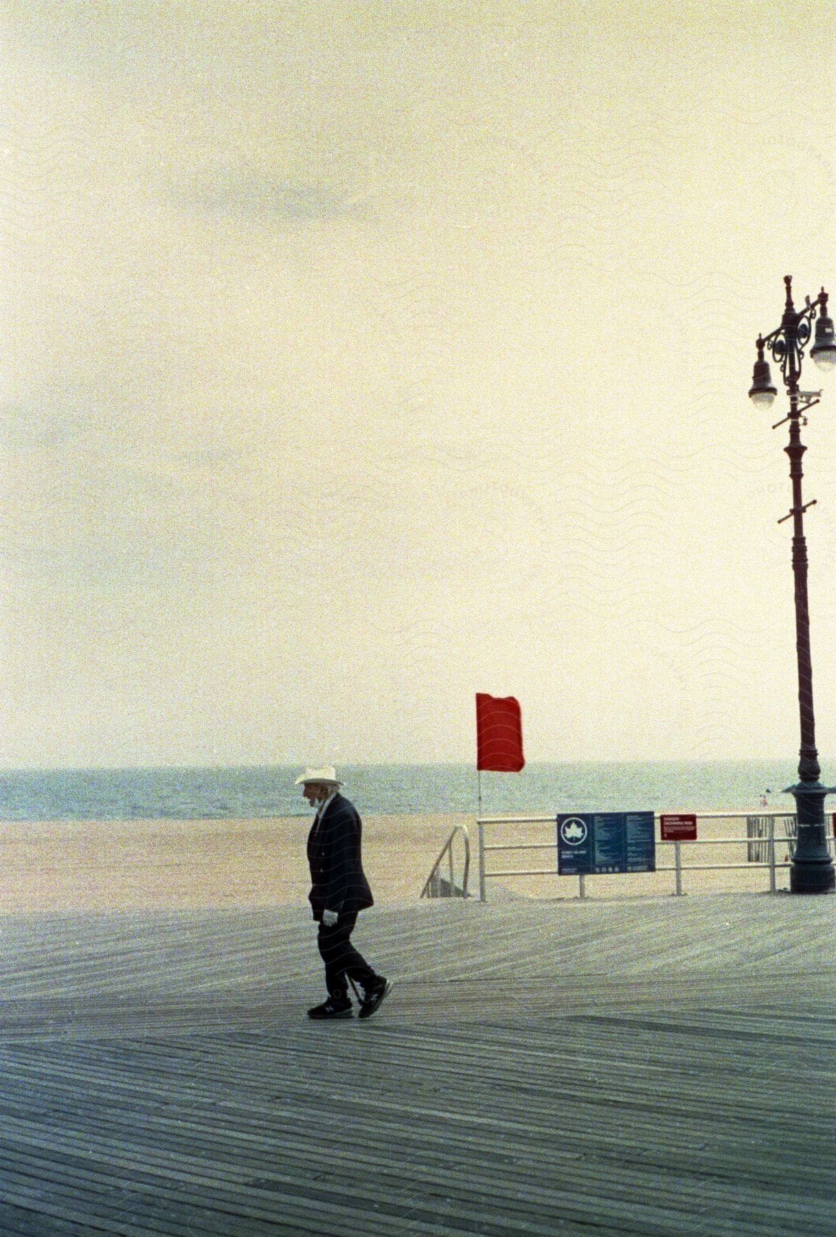 Man with white sun hat black blazer jacket pants and shoes walks along boardwalk near the beach at coney island