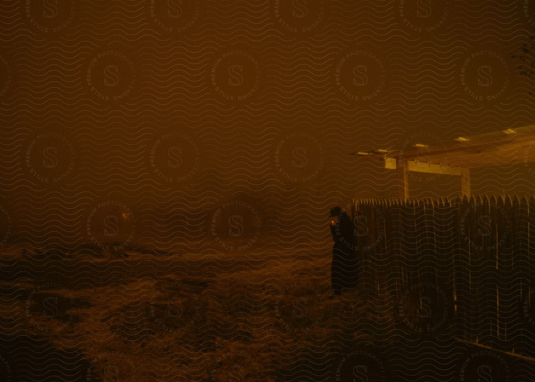 Man wearing black clothing smoking a cigarette while standing next to a fence at night