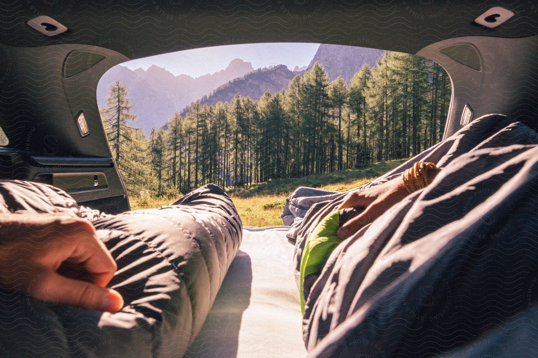 Couple In Sleeping Bags Looking Out At Forest From Back Of Vehicle On Sunny Day During Road Trip