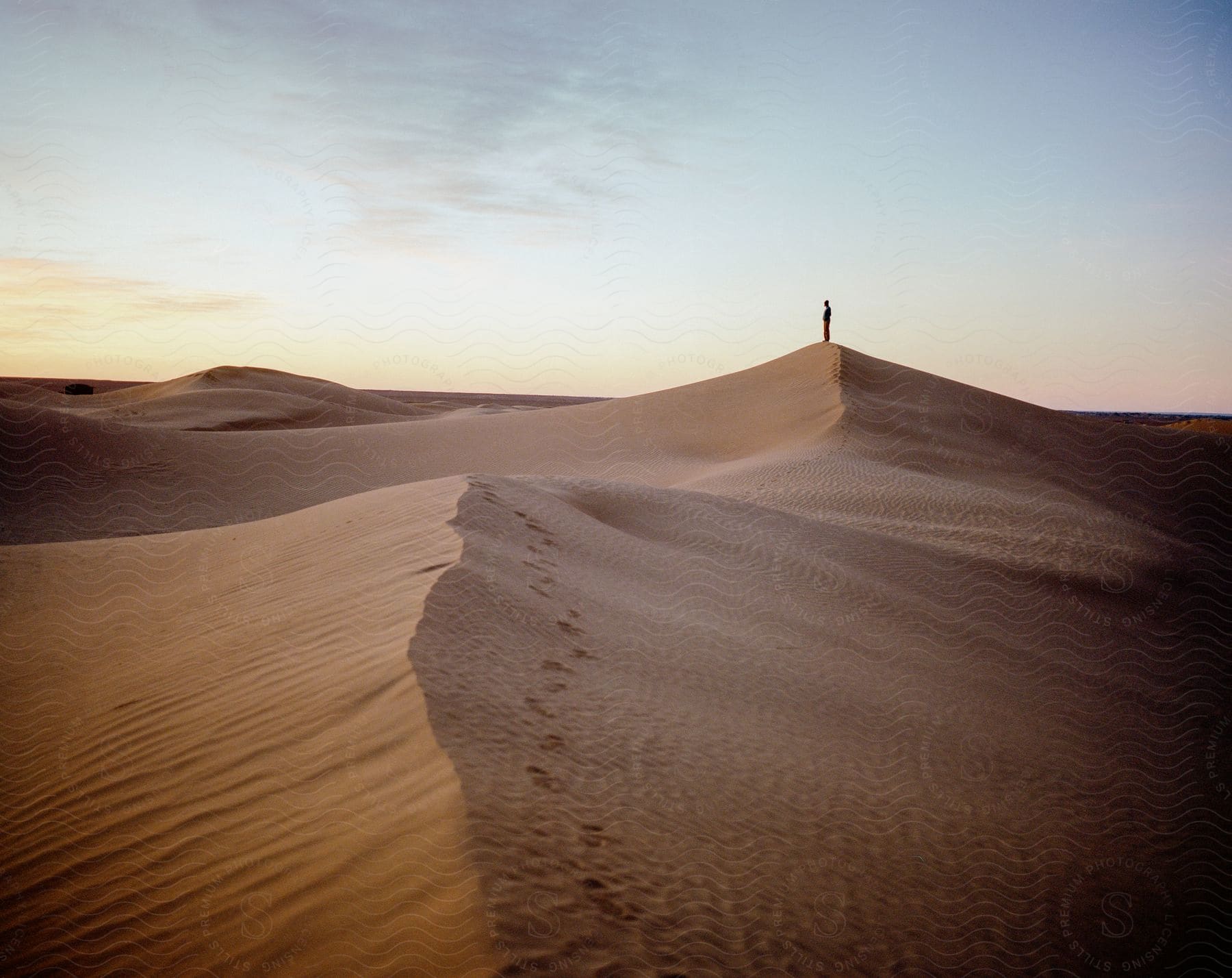 Person standing atop a mountain sand dune in the desert at sunset