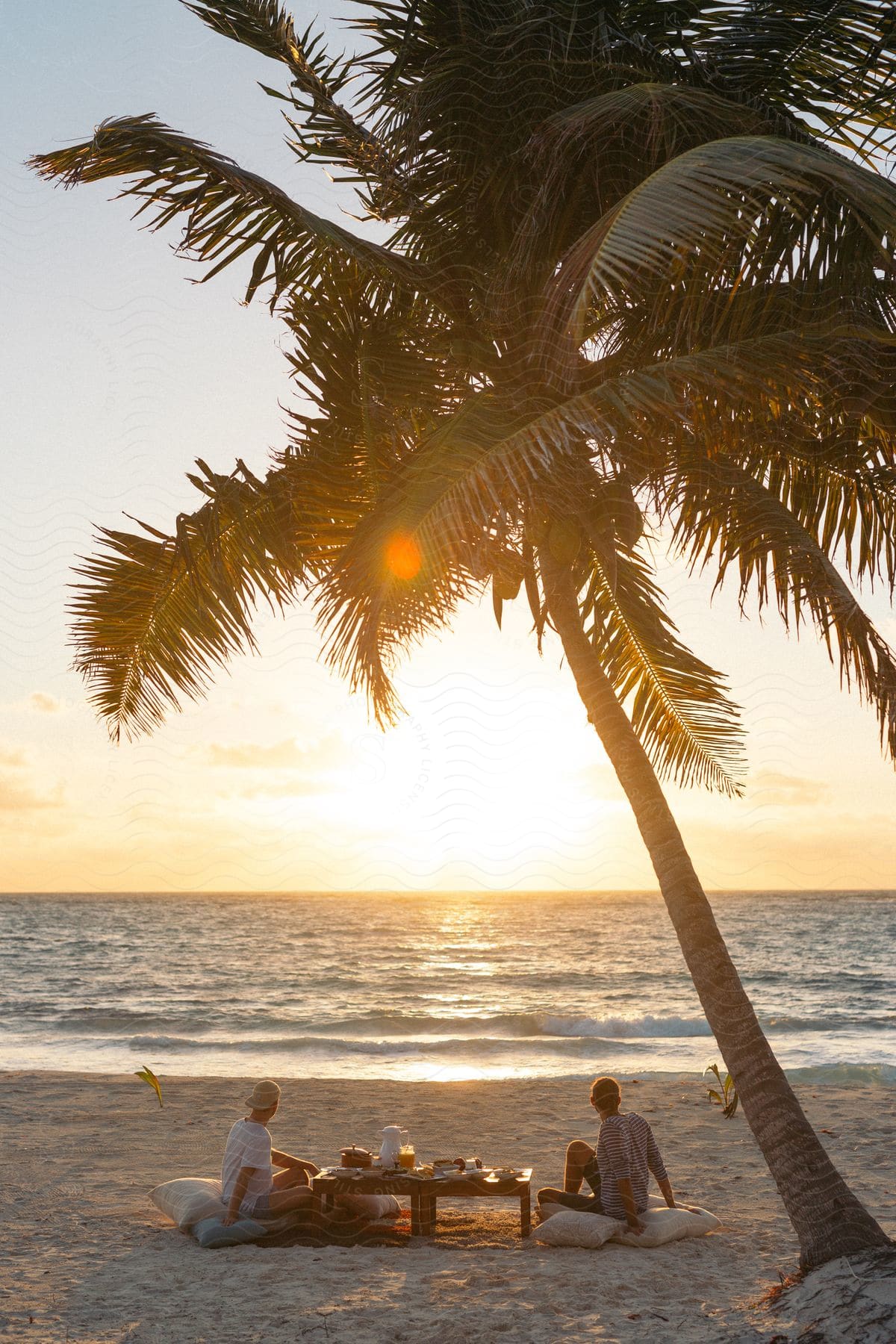 Two men sitting on pillows on the beach watch sunset over the sea next to palm tree