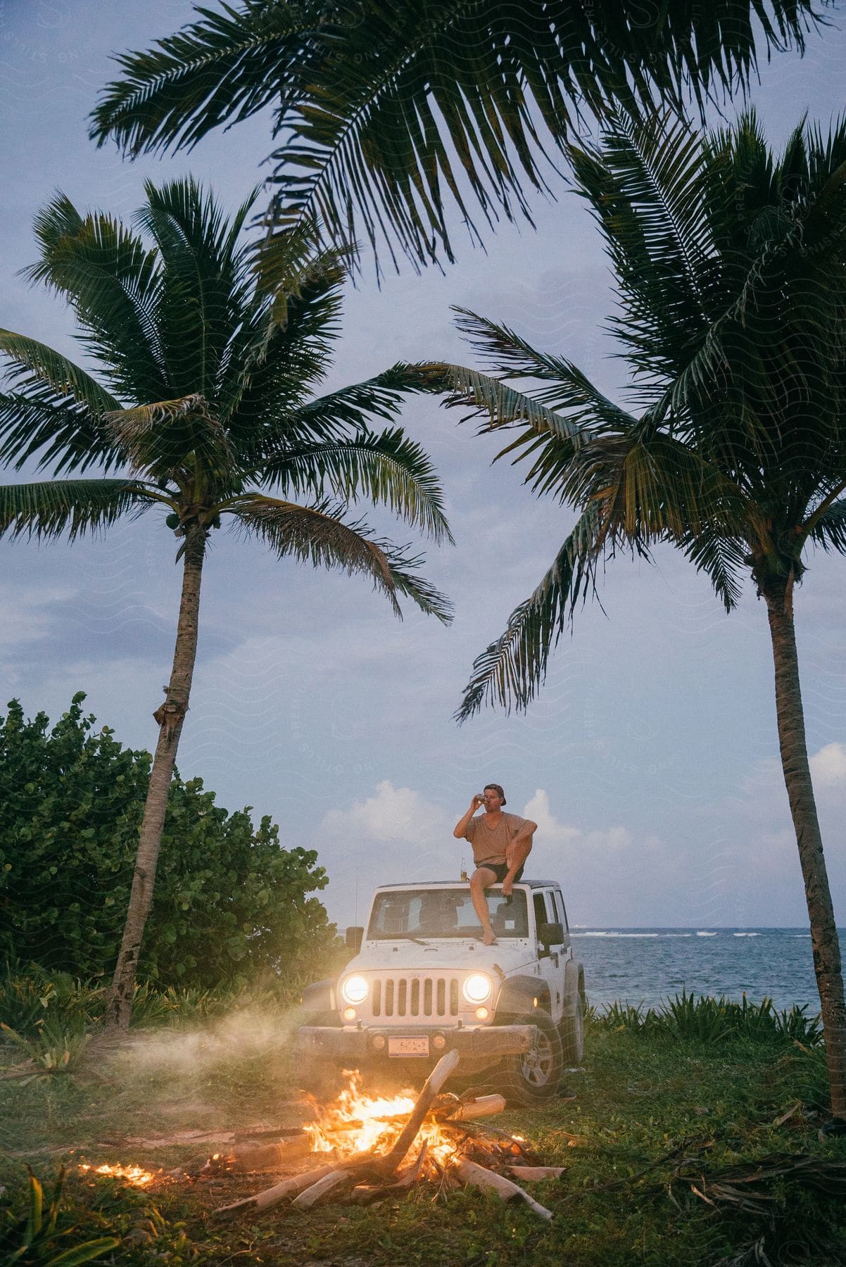 A man sitting on top of a parked car on the edge of the beach with a bonfire going