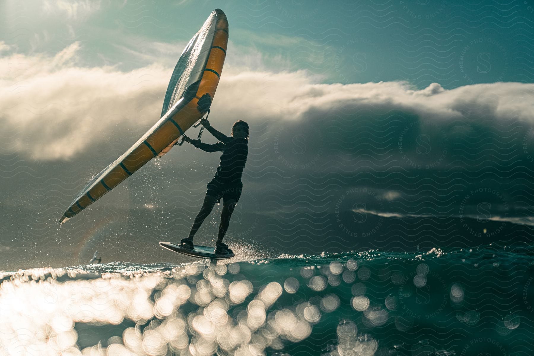 A woman windsurfs through ocean waves with storm clouds over a mountain range in the distance