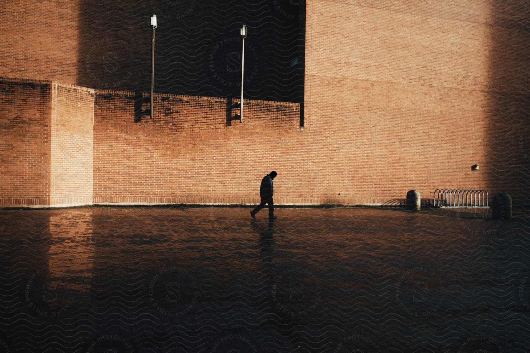 A person walking across wet pavement near a brick building at dusk or dawn