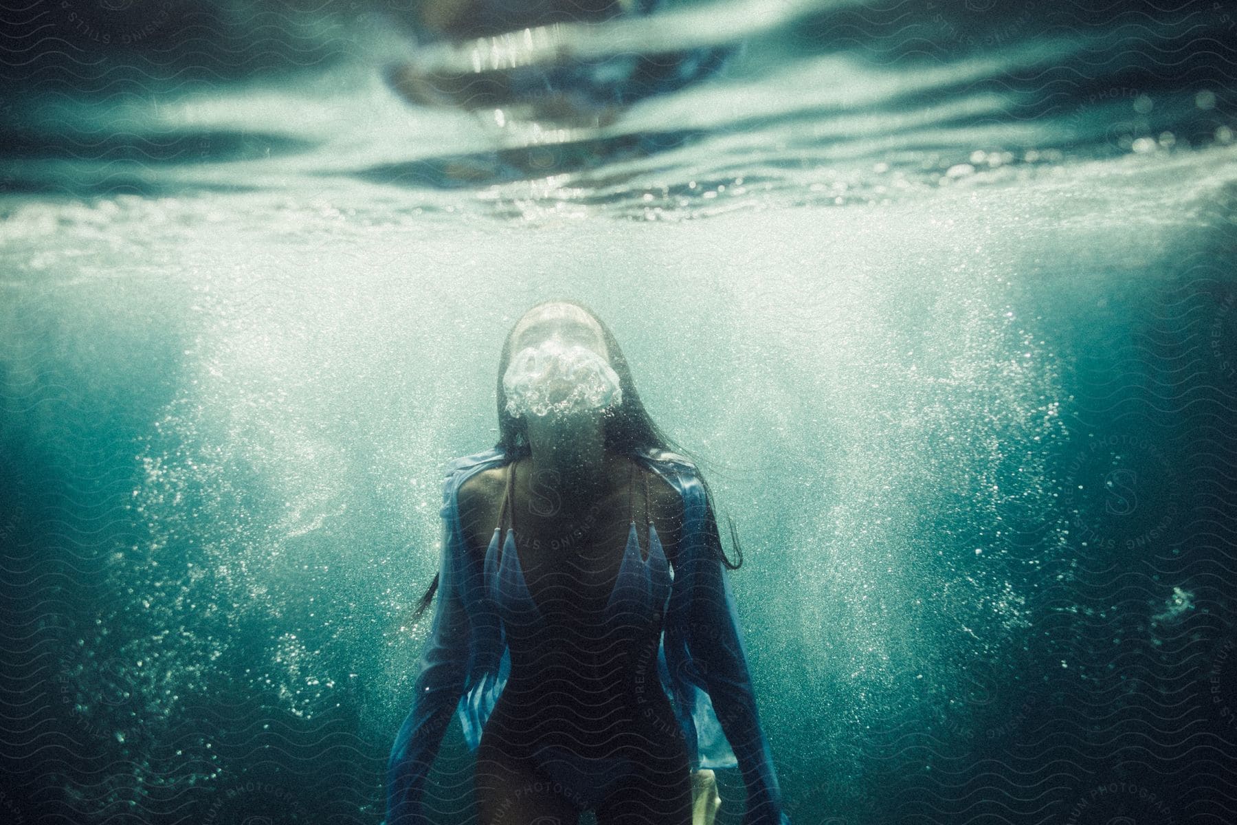 Dark haired woman dressed in blue bikini and shirt swimming and exhaling underwater in a body of water