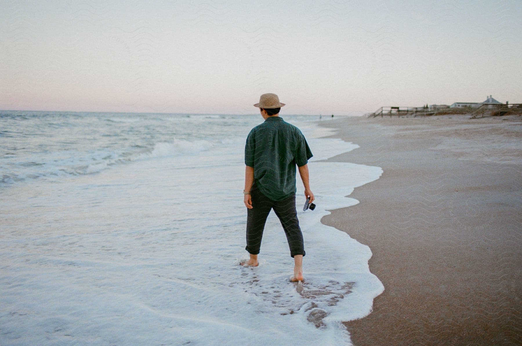 Man wearing a hat walking on a beach shoreline carrying a camera