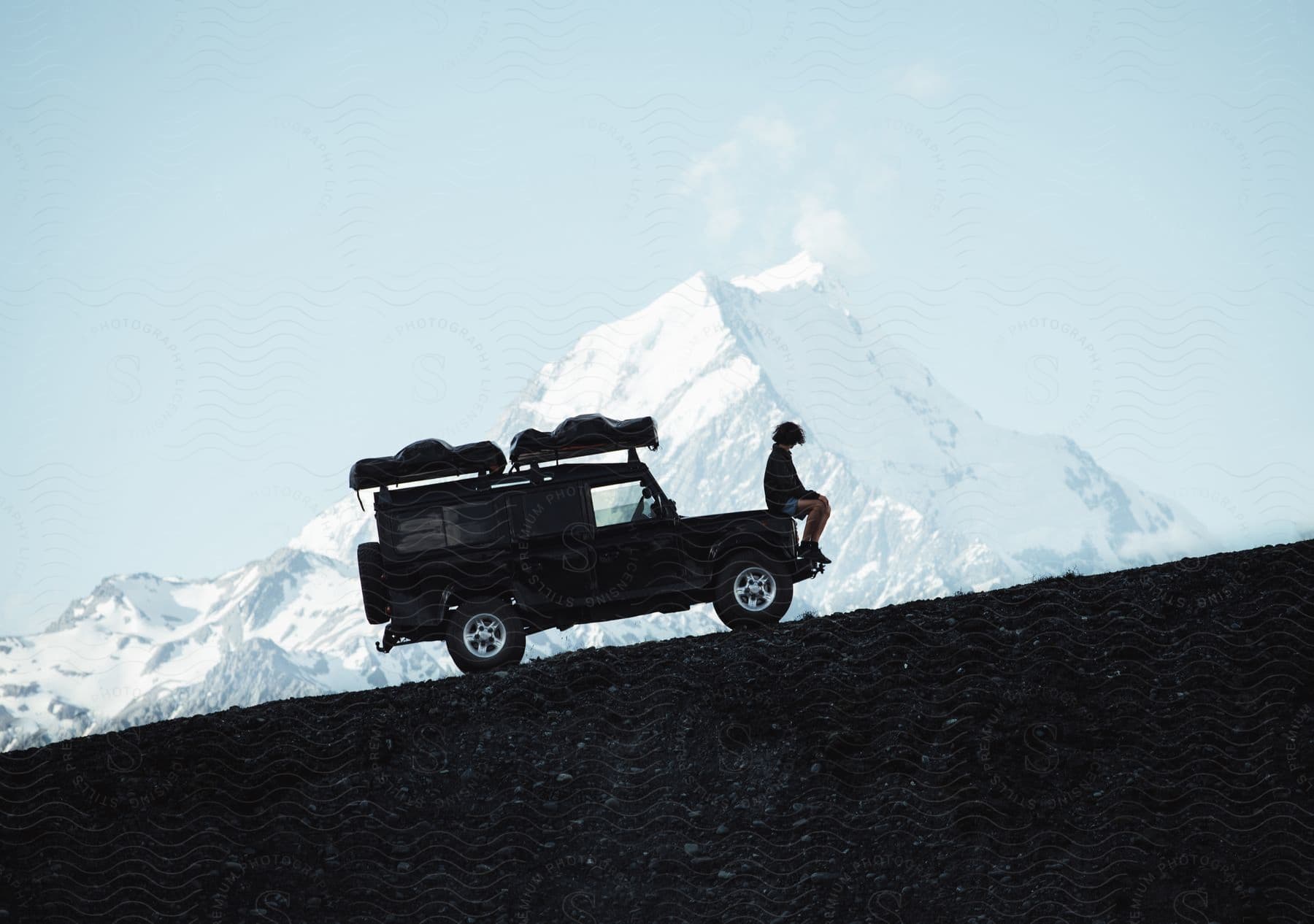 A man sits on a jeep parked on a snowy slope with mountains in the distance