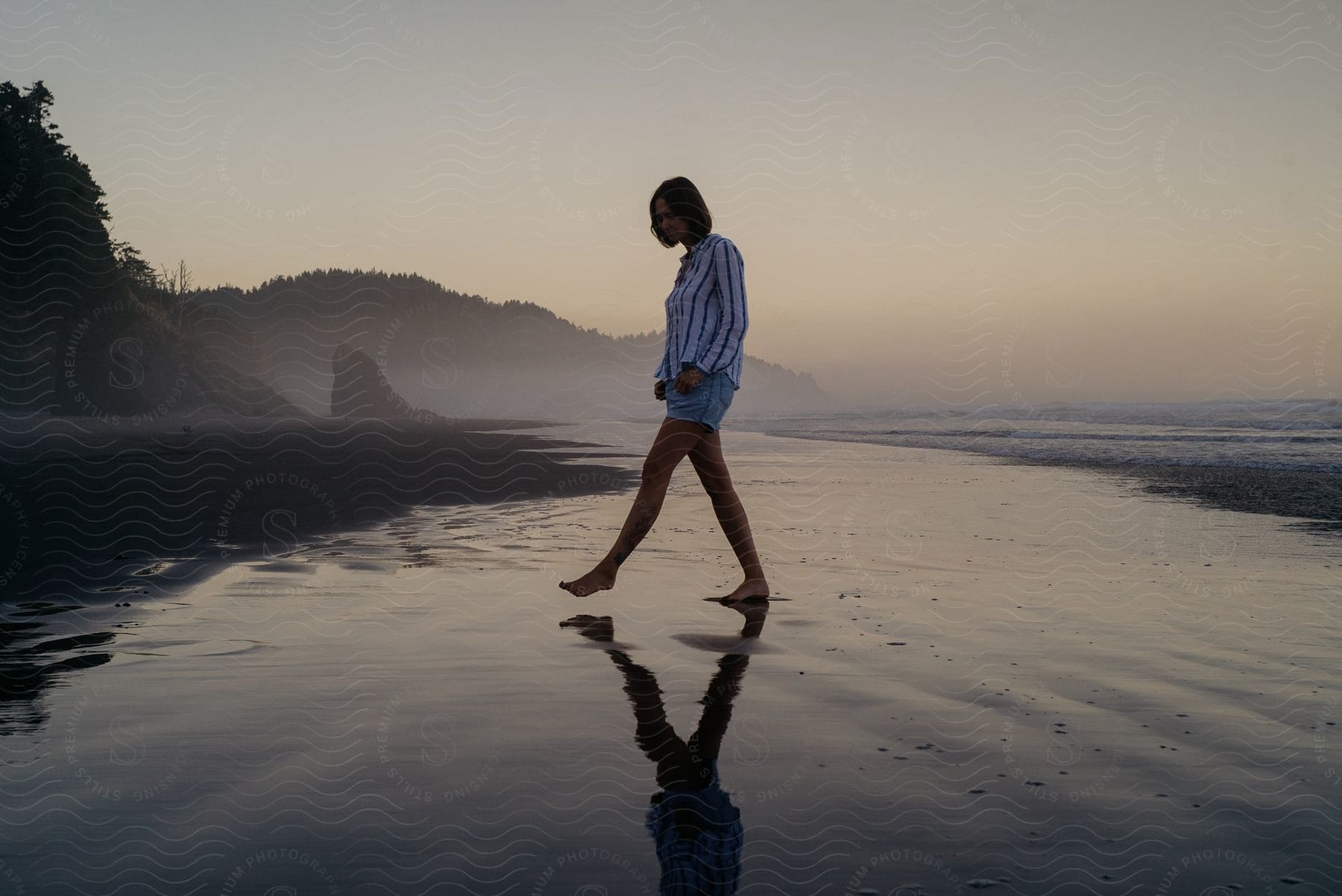 A woman standing close to the beach wearing shorts and a shirt with trees in the background