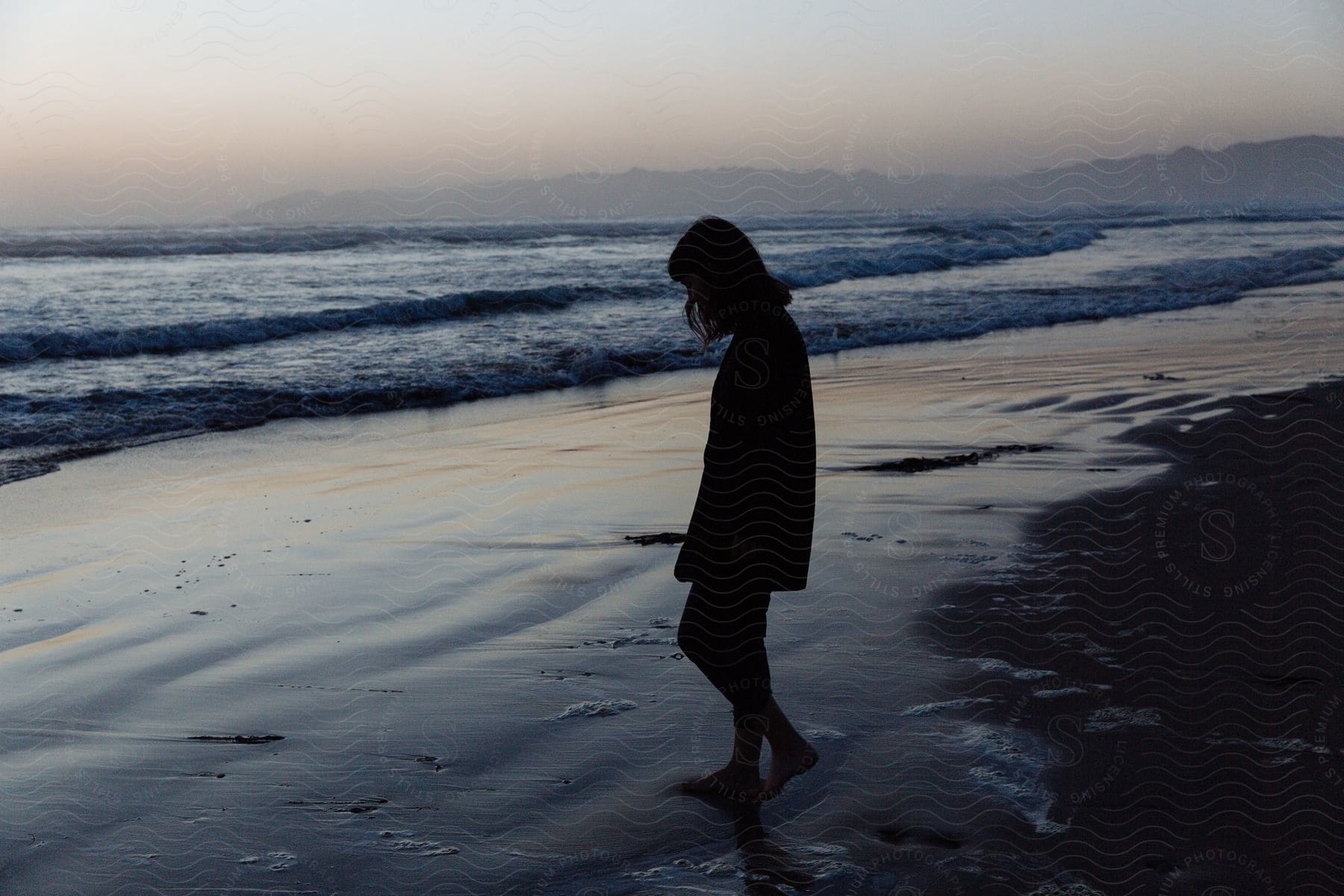 Silhouette of woman walking barefoot on beach with waves rolling into shore mountains in distance under hazy sky