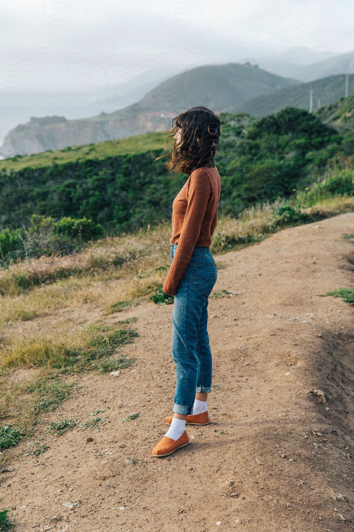 Woman with long dark hair standing on a hillside trail with mountains in the distance