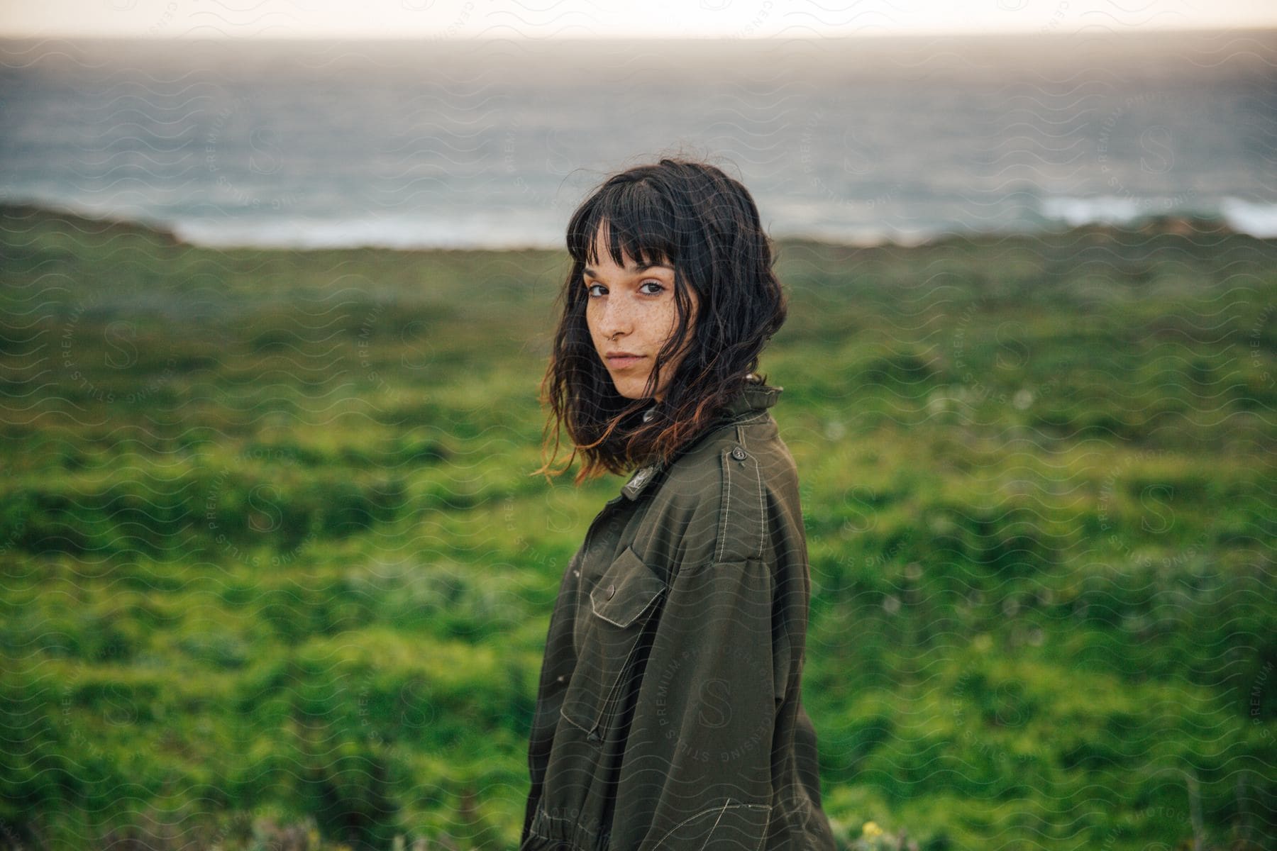 A person with black hair wearing a coat stands in a sunny field looking happy