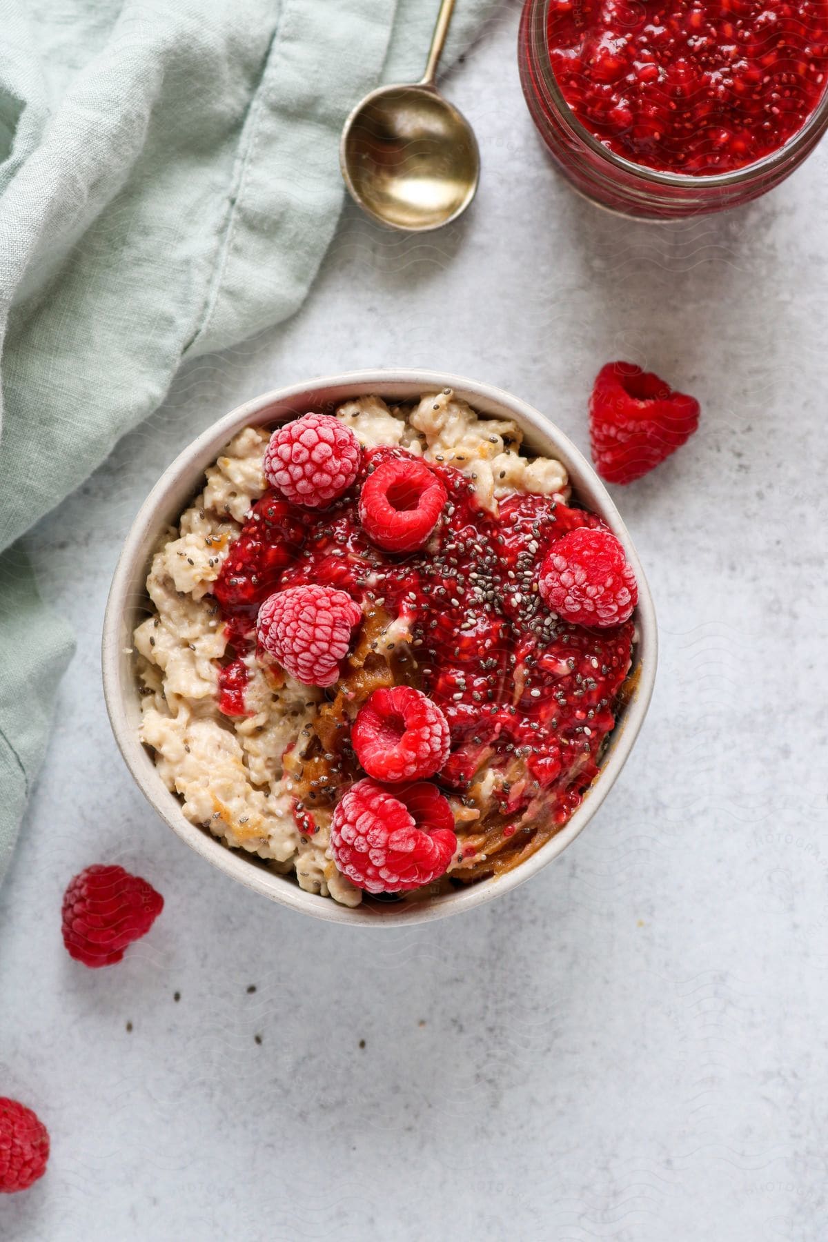 A bowl of oatmeal and fresh raspberries a jar of raspberry jam and a silver spoon on a white table