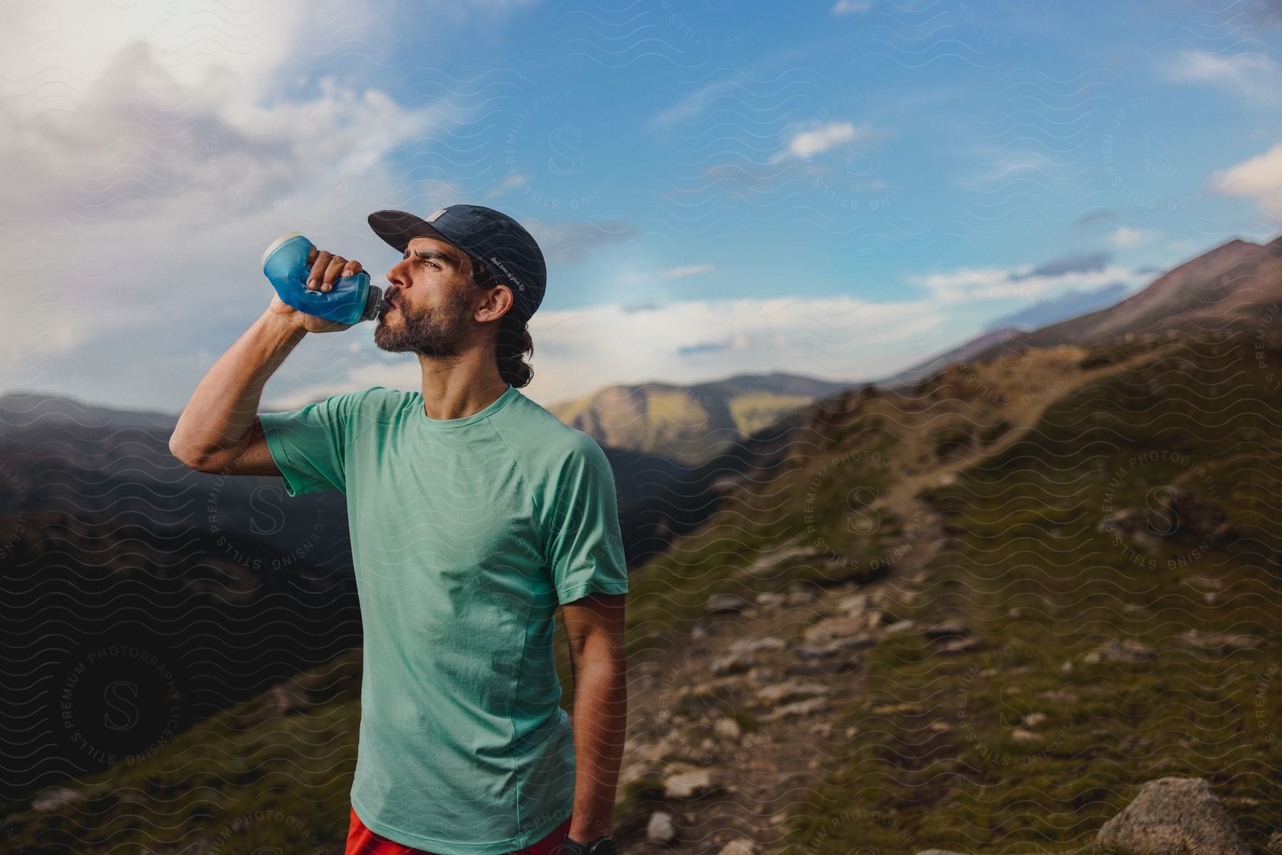 A man drinks from a water bottle while standing on a mountain under a cloudy sky