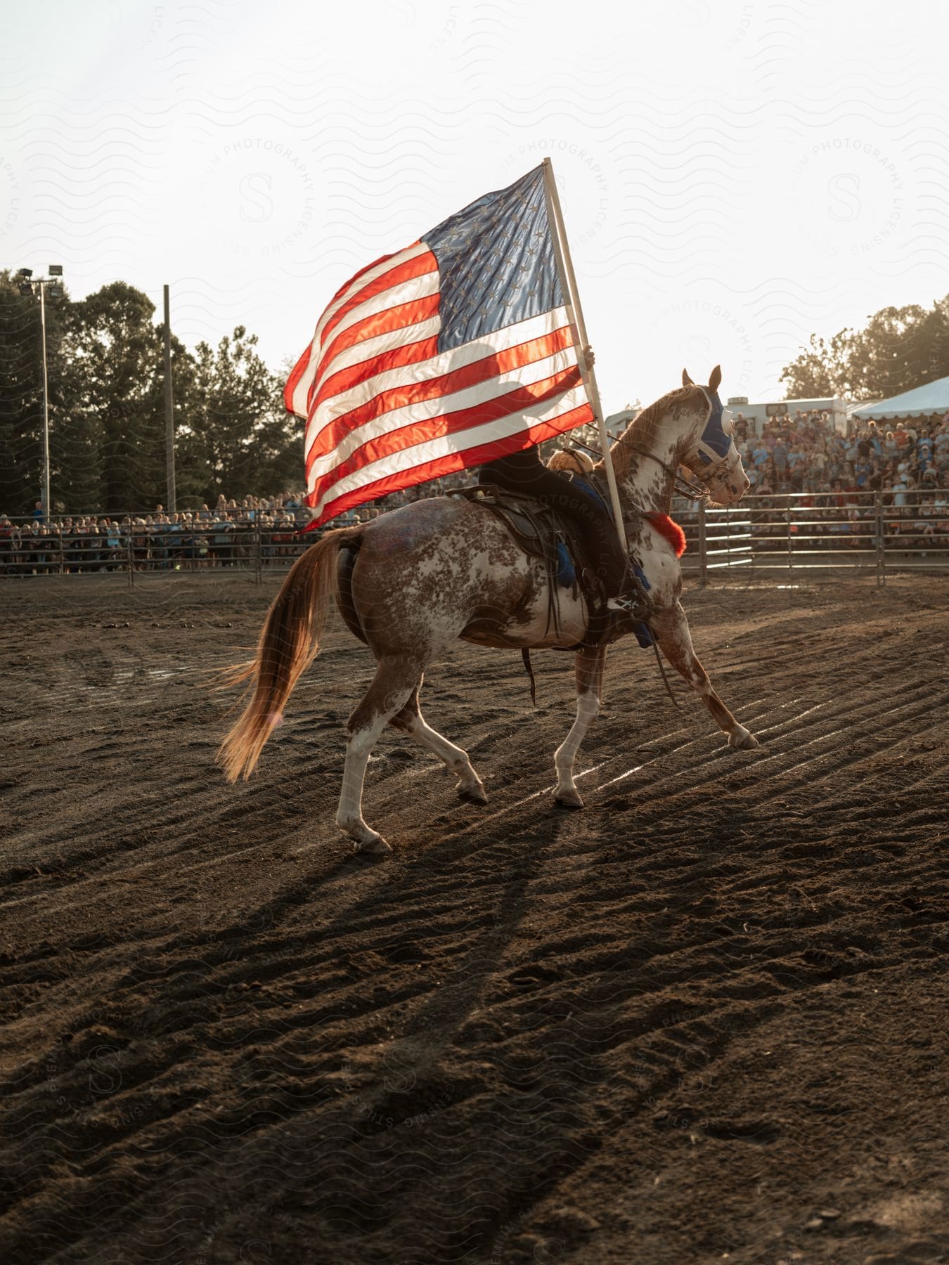 A large crowd gathers in a sports field as a determined person on a horse achieves victory with the sky and a flag in the background