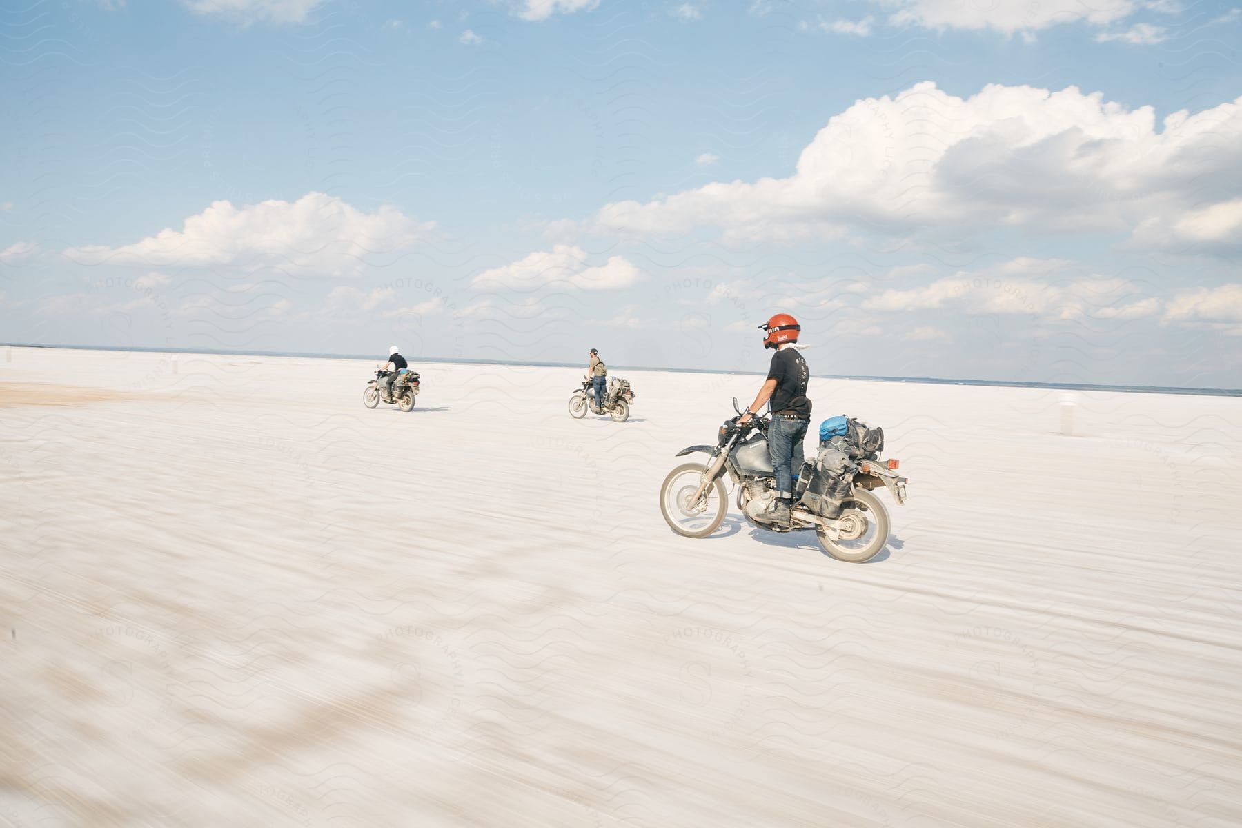 Three men riding motorcycles at full speed in a large desert during the daytime