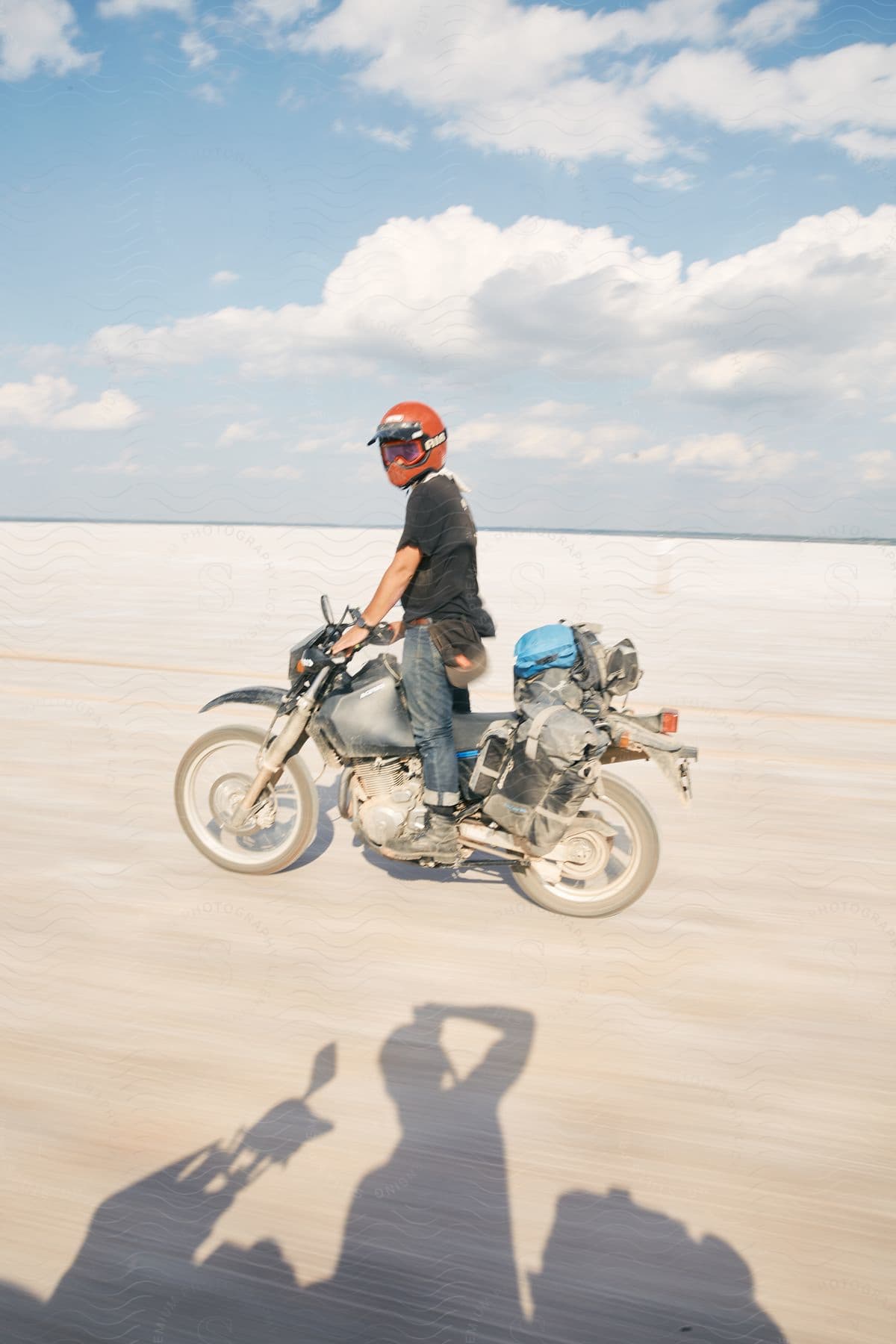 A man confidently rides his motorcycle amidst a vast expanse of dry land with white sand stretching as far as the eye can see