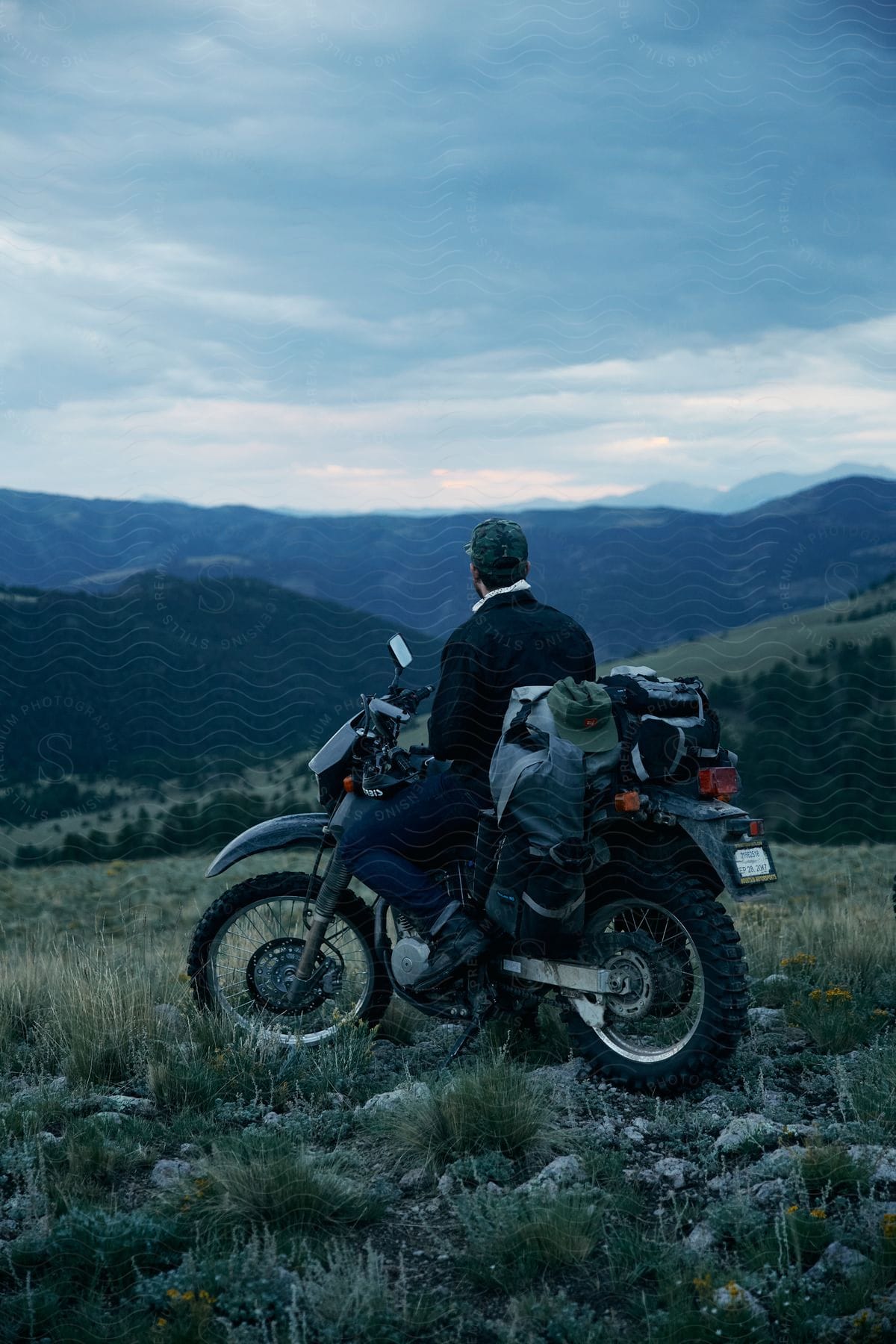 A man sits on his motorcycle in a field looking at mountains under a cloudy sky