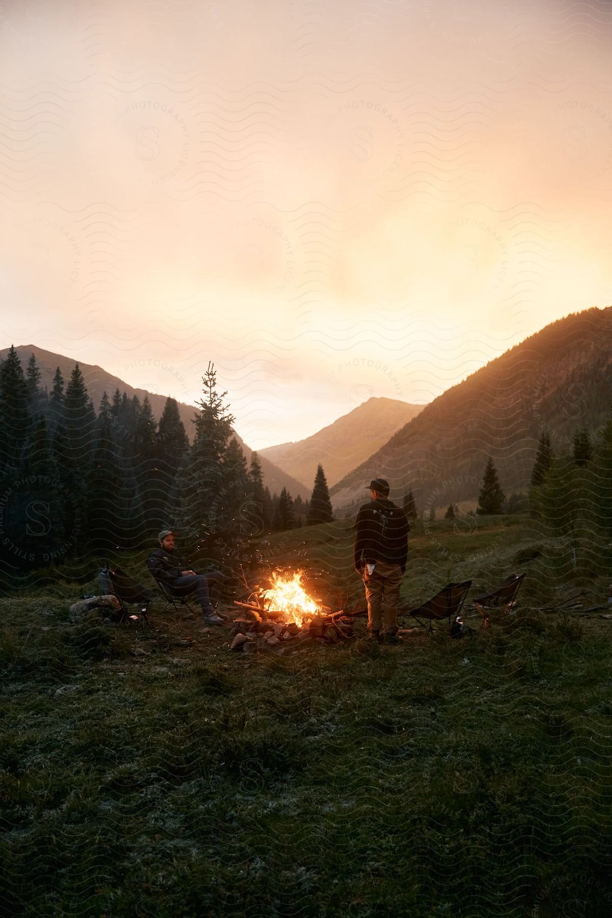 Two people sit by a bonfire in the wilderness surrounded by mountains and vegetation under a colorful sunset sky