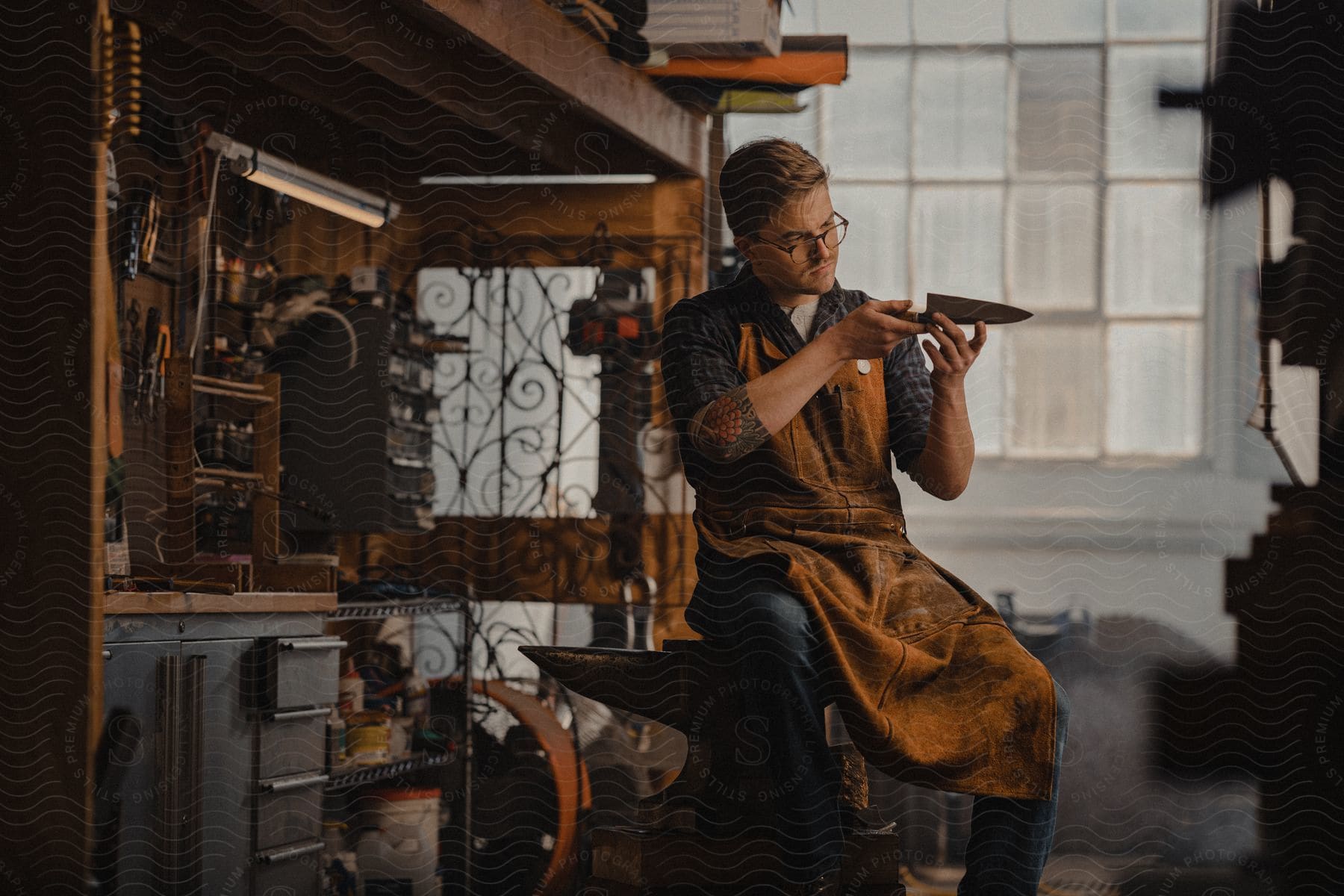 A man inspects a forged metal knife blade while sitting on an anvil in a rustcovered apron in an oldfashioned metal shop