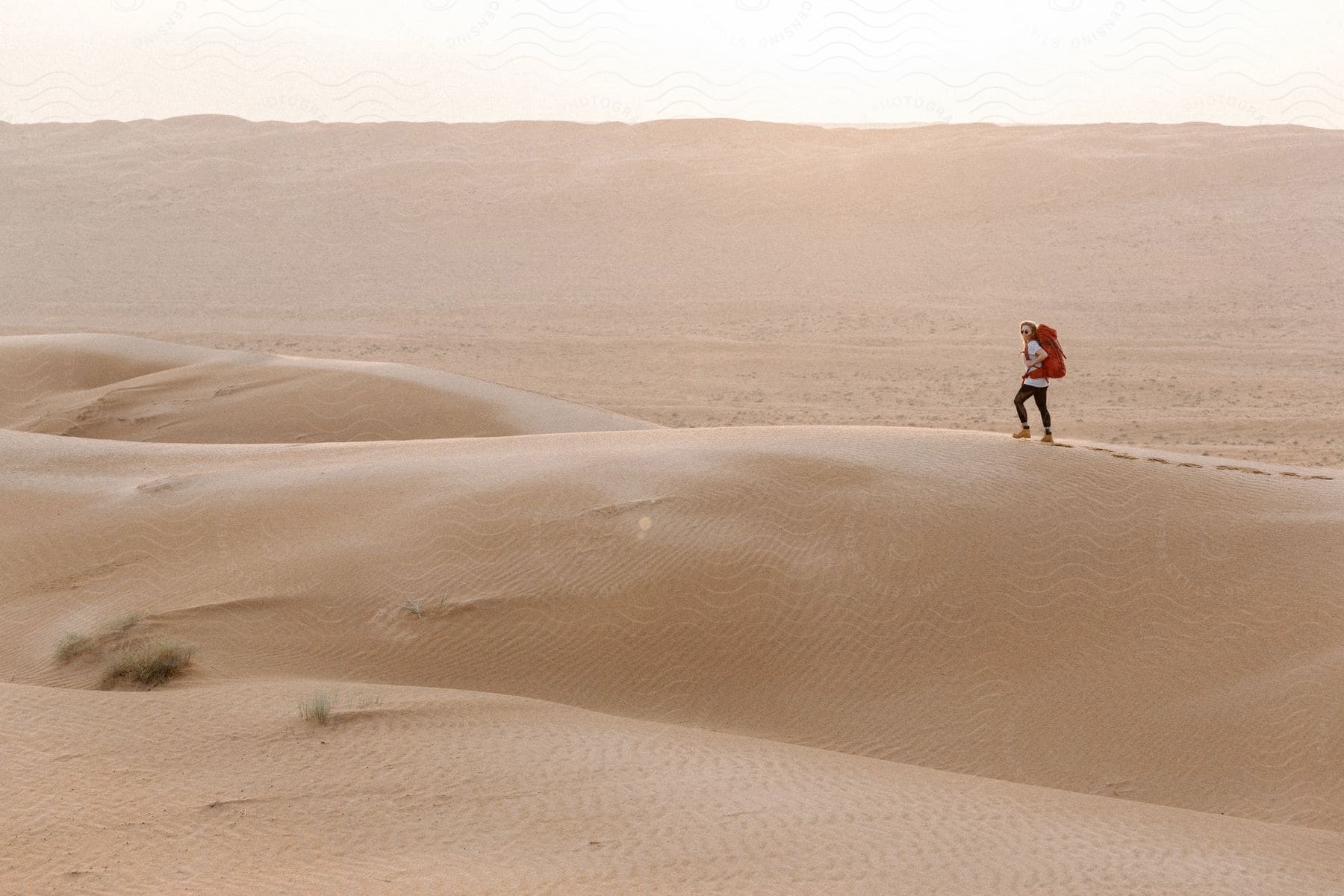 A hiker with a red backpack walks across a desert hill leaving footprints in the sand