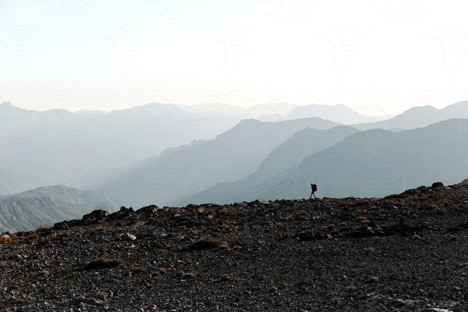 A Person Walks On Rocky Terrain On Top Of A Mountain In A Mountain Range On A Bright Day