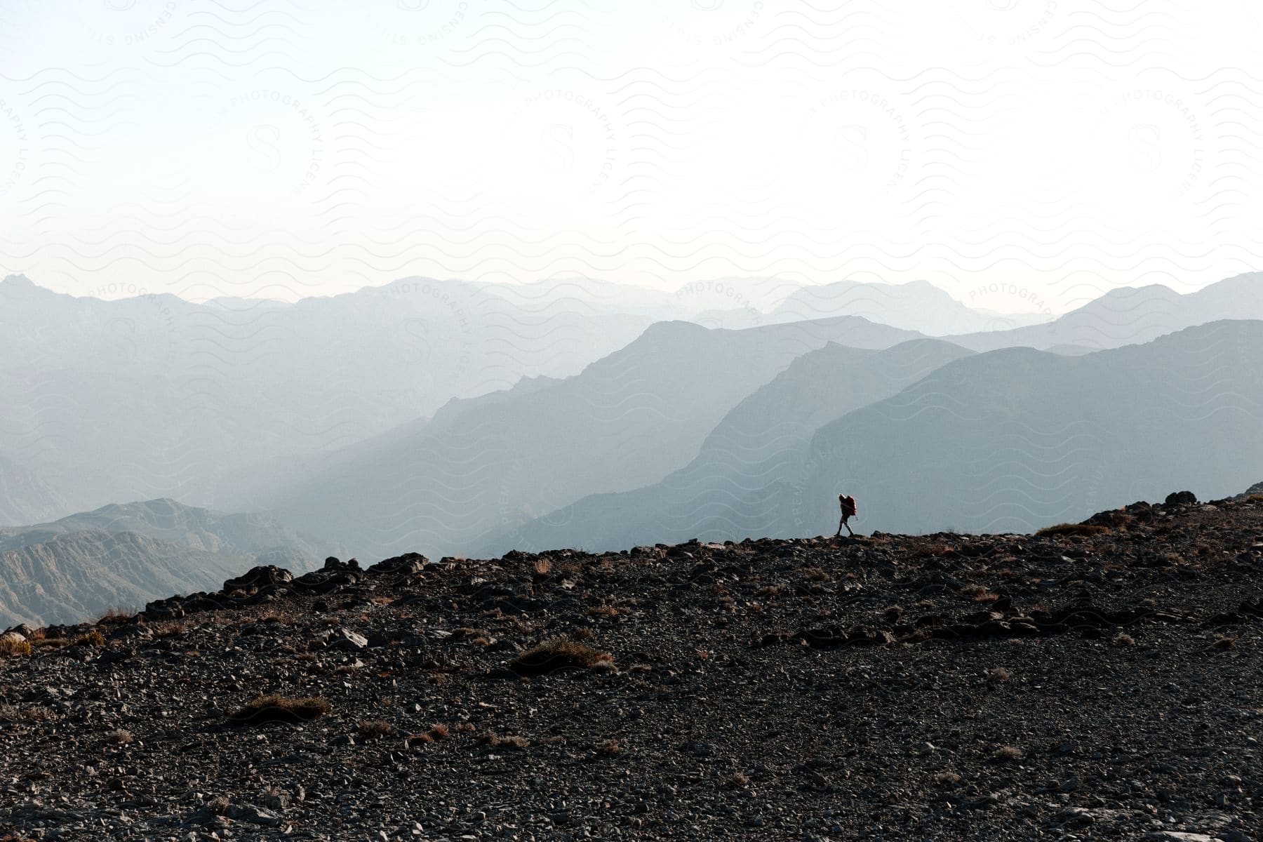 A person walks on rocky terrain on top of a mountain in a mountain range on a bright day