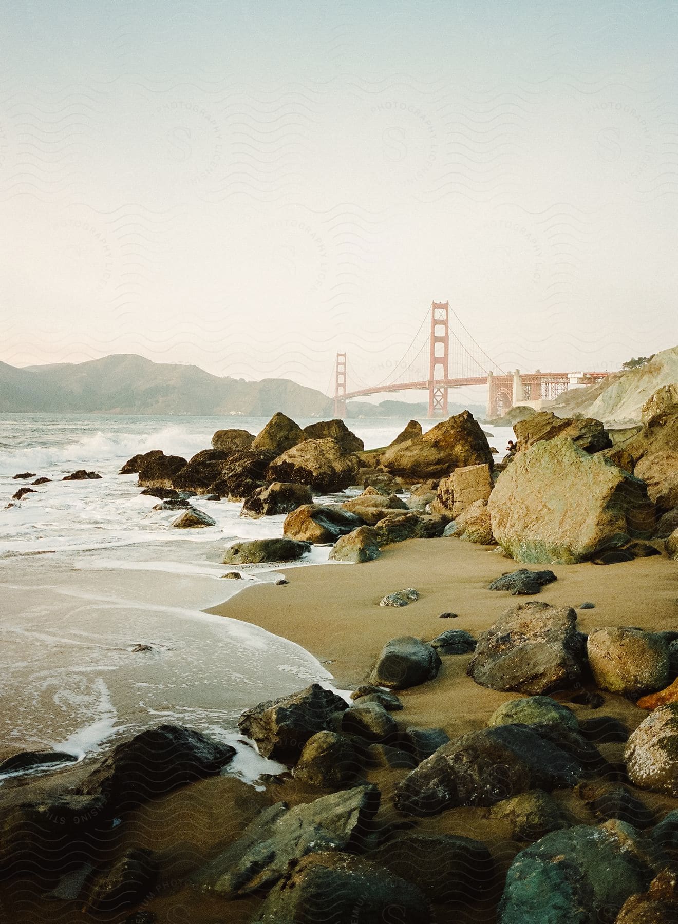 A foggy day on a rocky shoreline with water starting to wash up near a large bridge