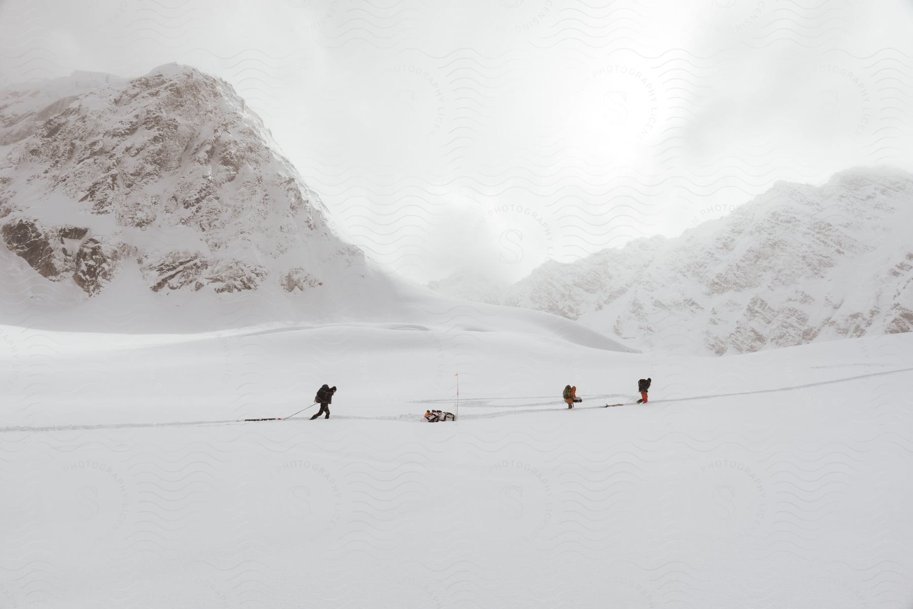 Three people with backpacks and sleds crossing snowy terrain with mountains in the distance under a white winter sky